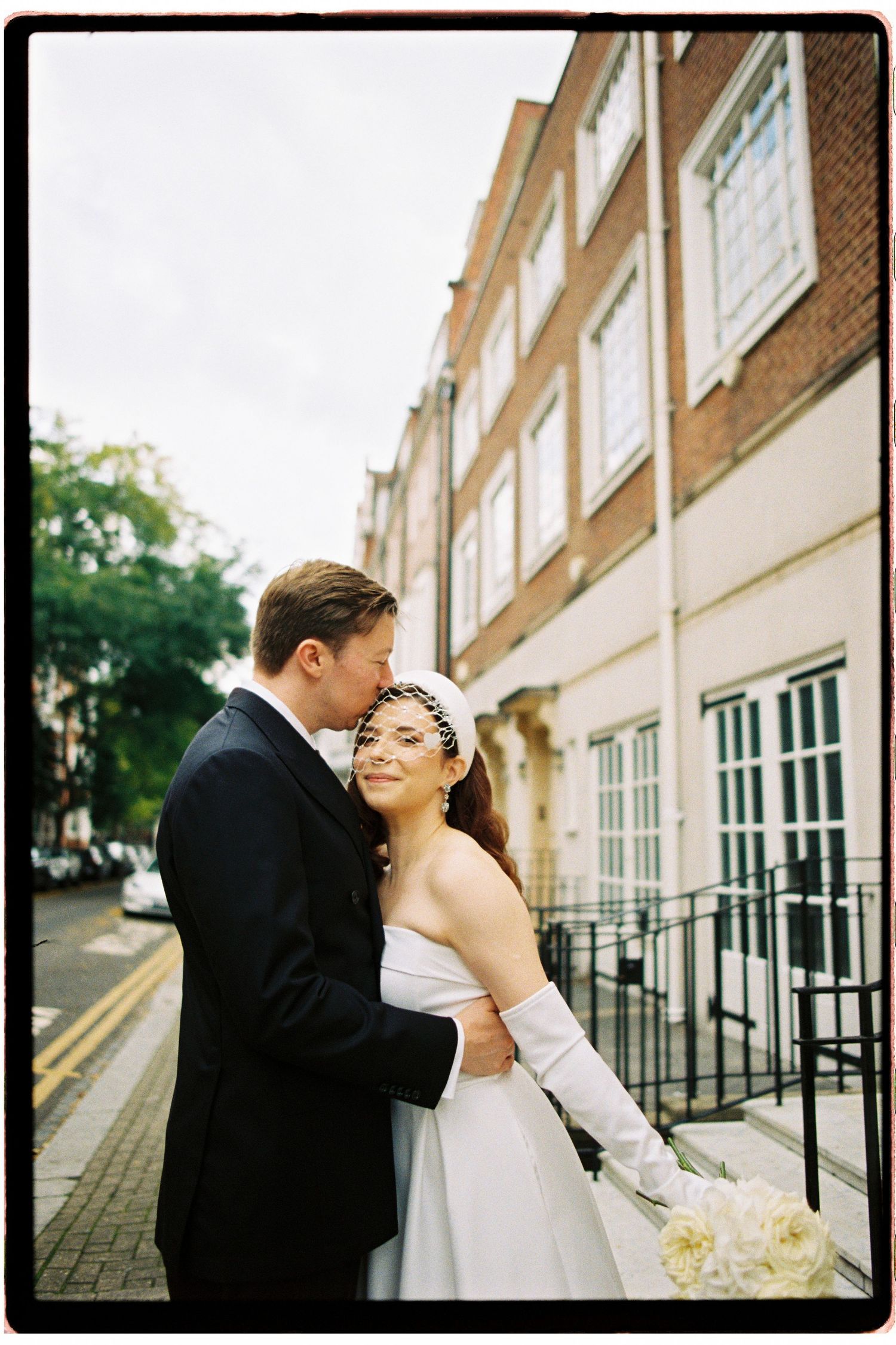 A bride and groom share a tender embrace on a London street, she wearing a white strapless gown and gloves holding a bouquet.