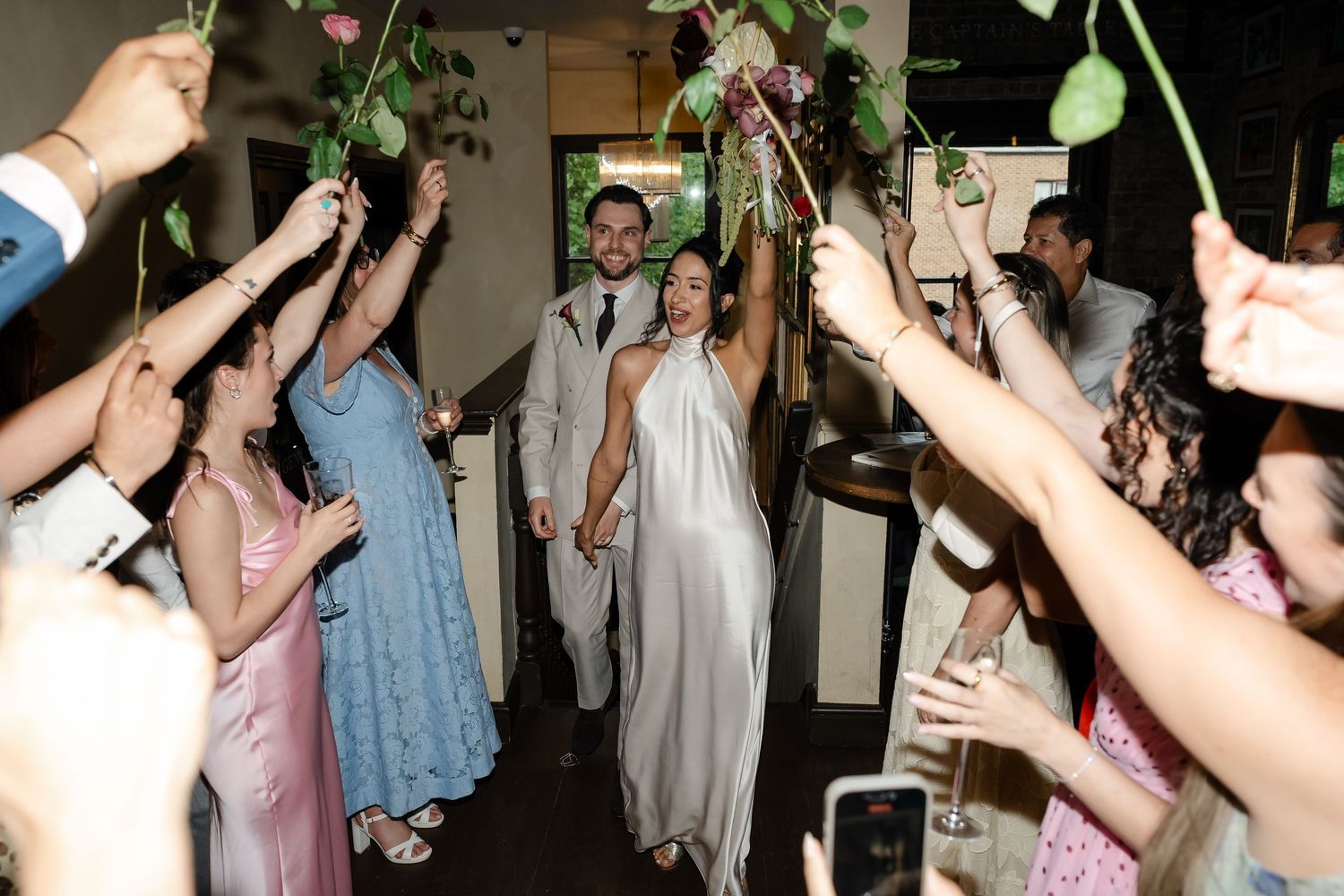 Newlyweds walk through a floral arch as wedding guests raise flowers in celebration.