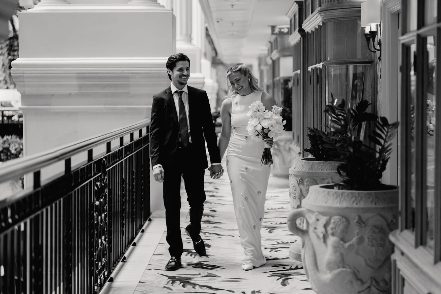 Bride and groom walking hand in hand along an elegant hotel corridor in a black and white photo.