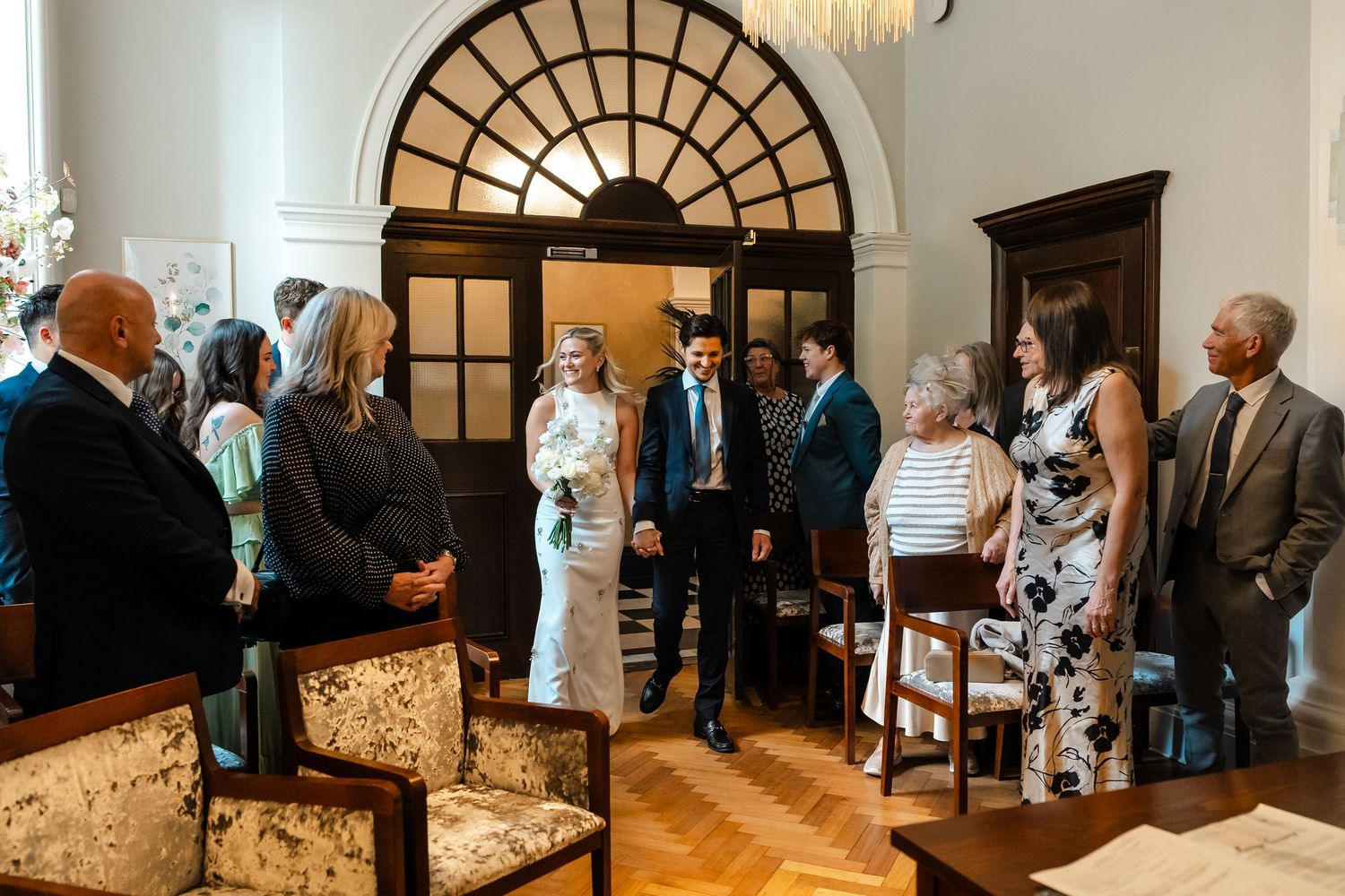 Wedding guests mingle in elegant ceremony room with arched windows and wooden parquet flooring.