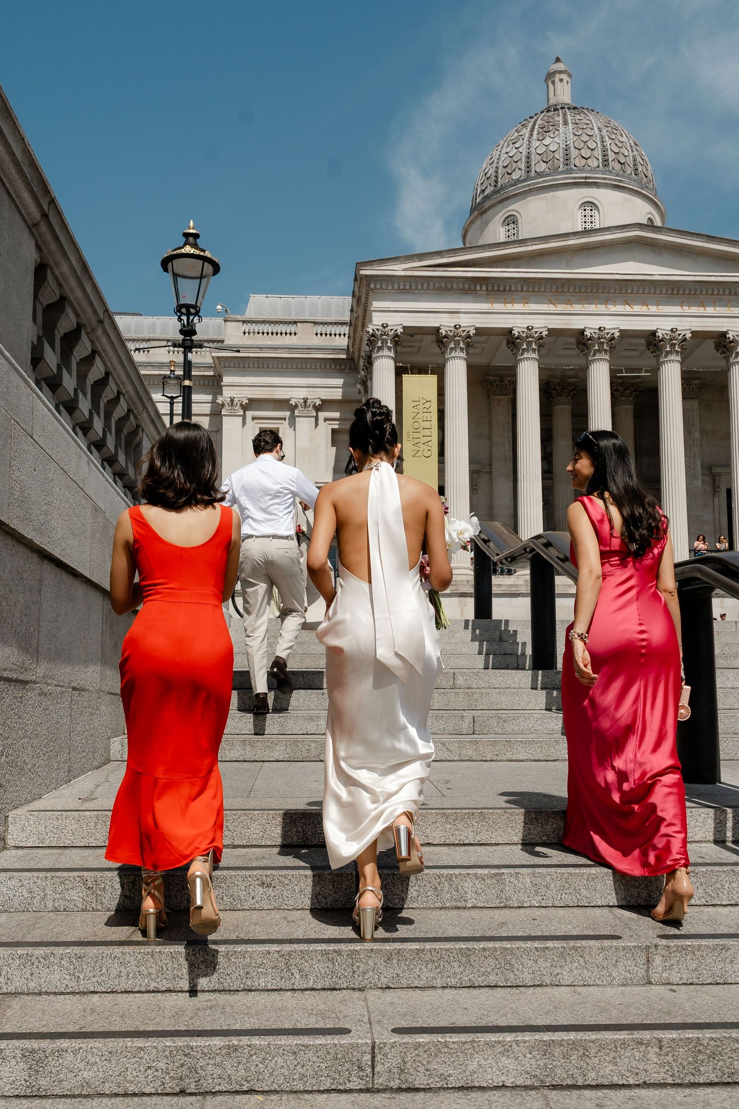Bride and two bridesmaids in red and pink gowns ascending steps toward the National Gallery in London.