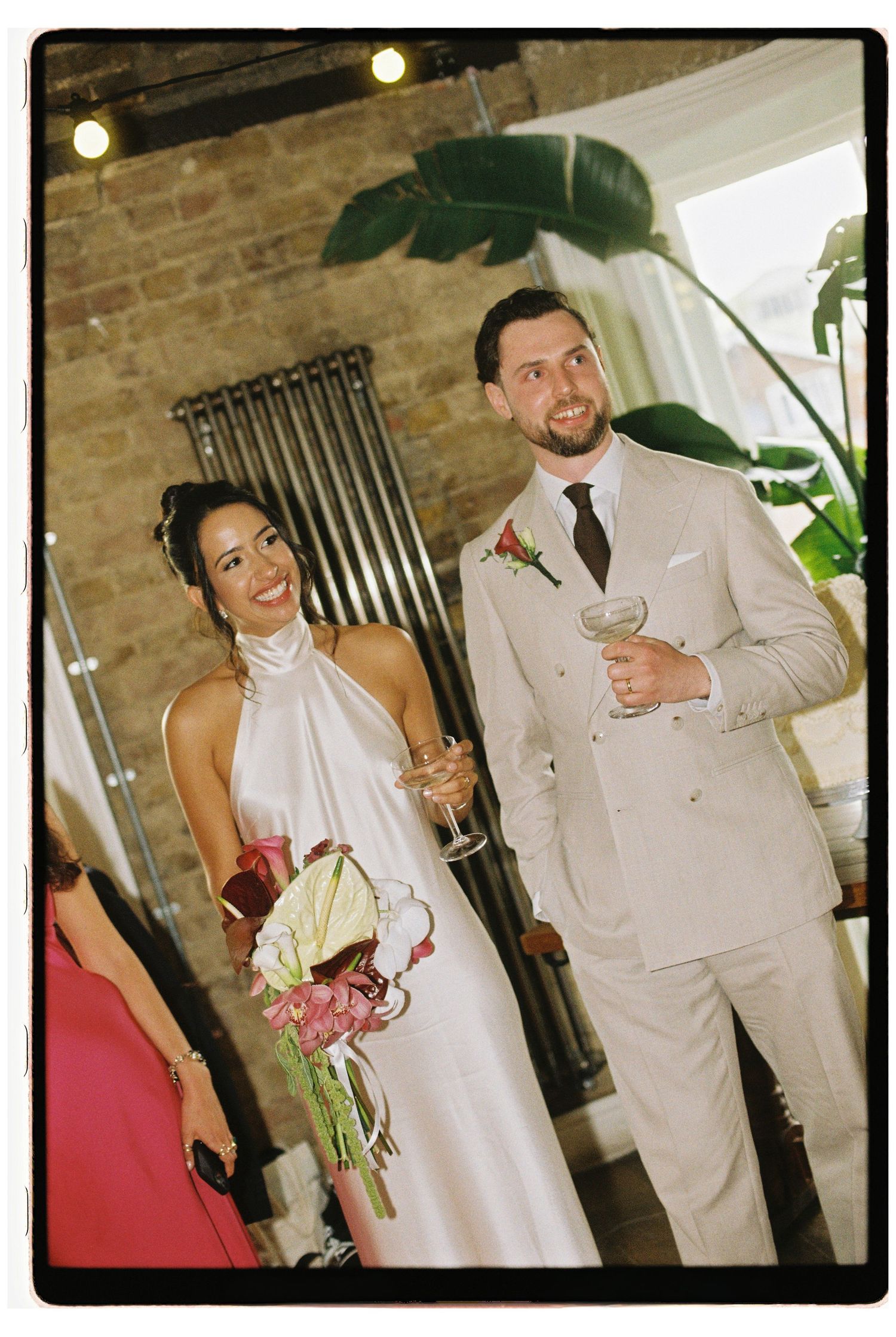 Bride and groom smiling holding champagne glasses in industrial brick venue with tropical plant decor.