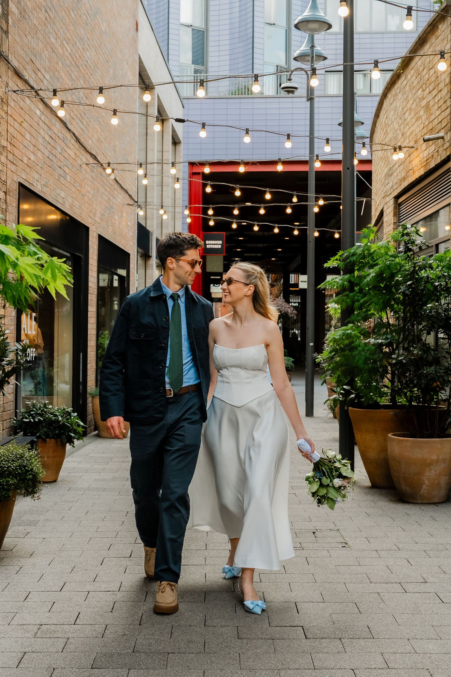 Bride and groom strolling through charming alley adorned with string lights and greenery.