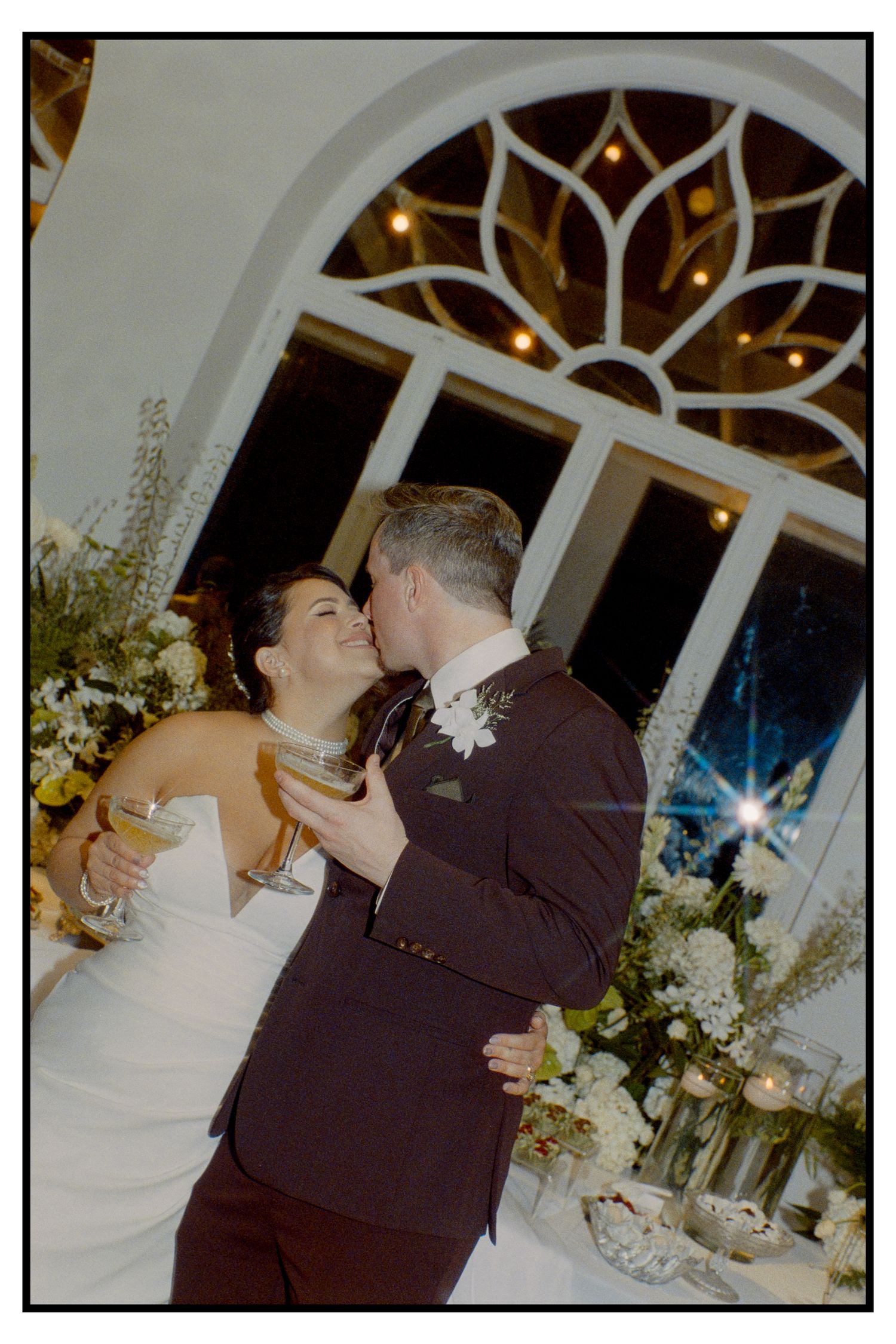 Newlyweds share a romantic kiss during wedding reception beneath elegant arched stained glass window.