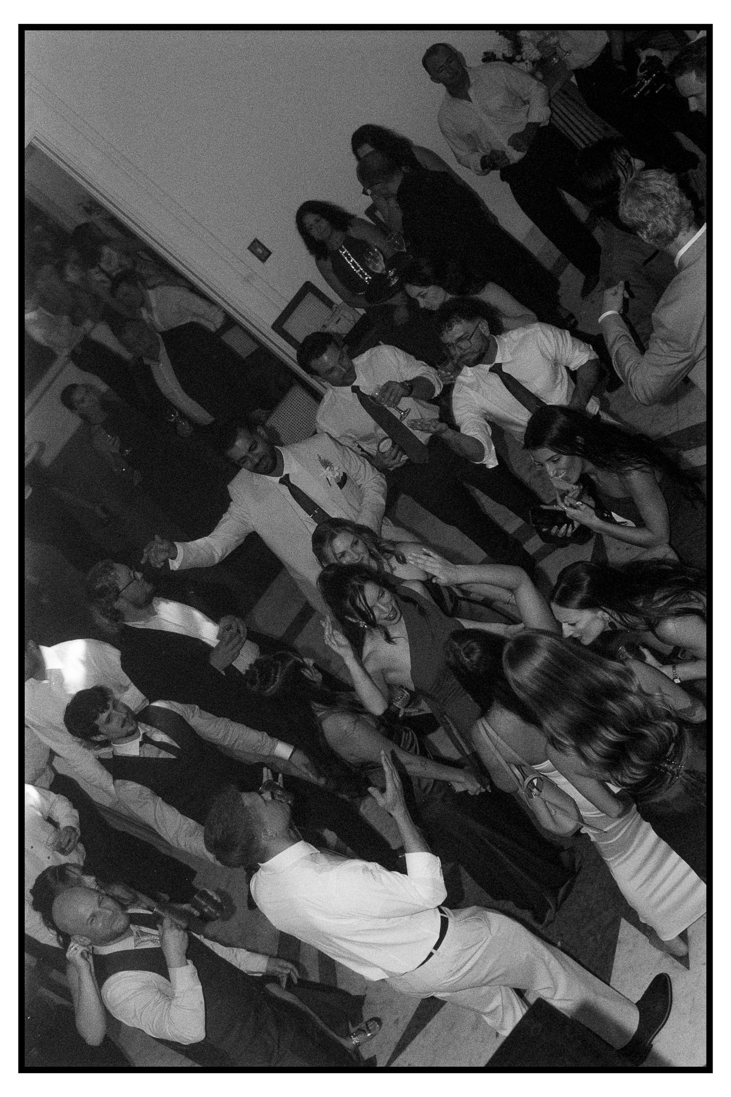 Black and white photo of elegantly dressed wedding guests mingling and dancing at a reception.