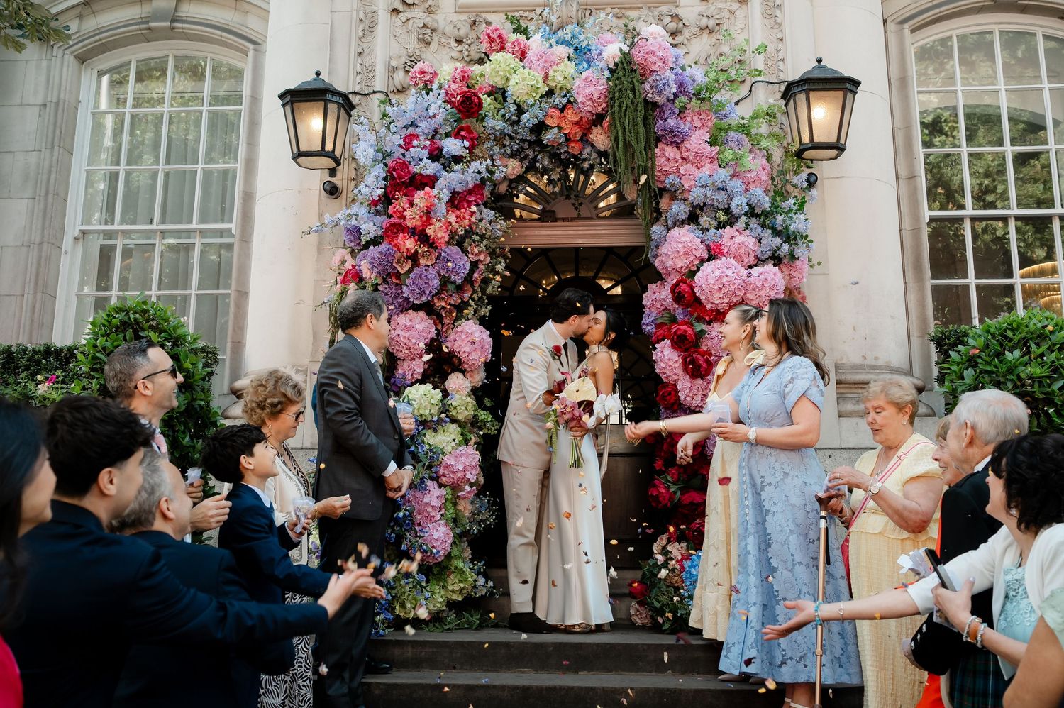 Newlyweds kissing under stunning colorful hydrangea floral arch as guests celebrate with confetti on steps.