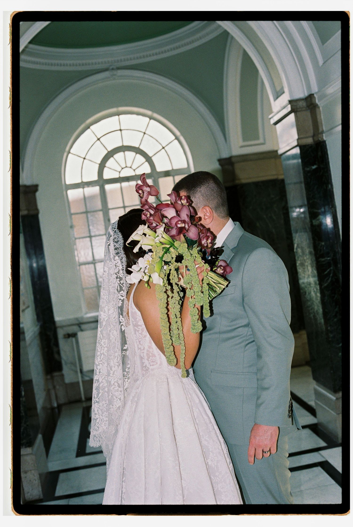 Bride and groom sharing a kiss in elegant venue, bride wearing backless lace gown with cascading floral bouquet.