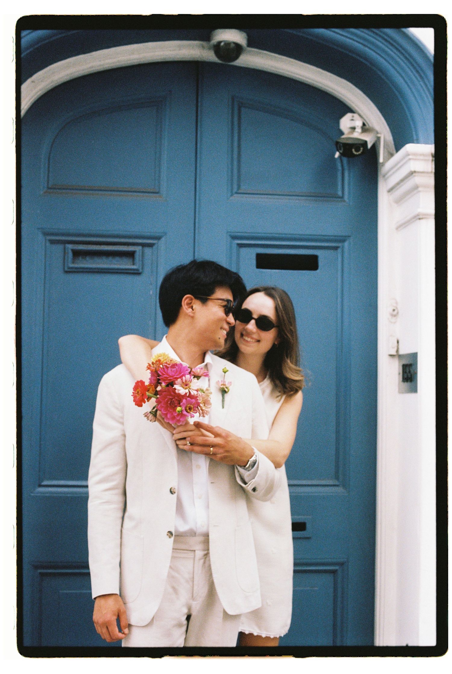 Couple in white attire embracing before a blue door, holding colorful flowers, in a film photography style.
