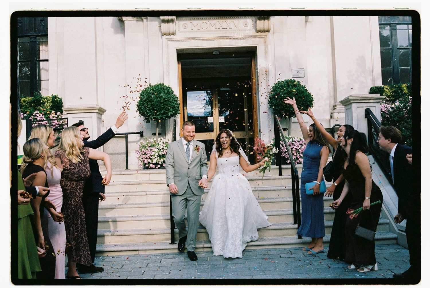Bride and groom exit venue steps as guests throw confetti during outdoor wedding celebration.