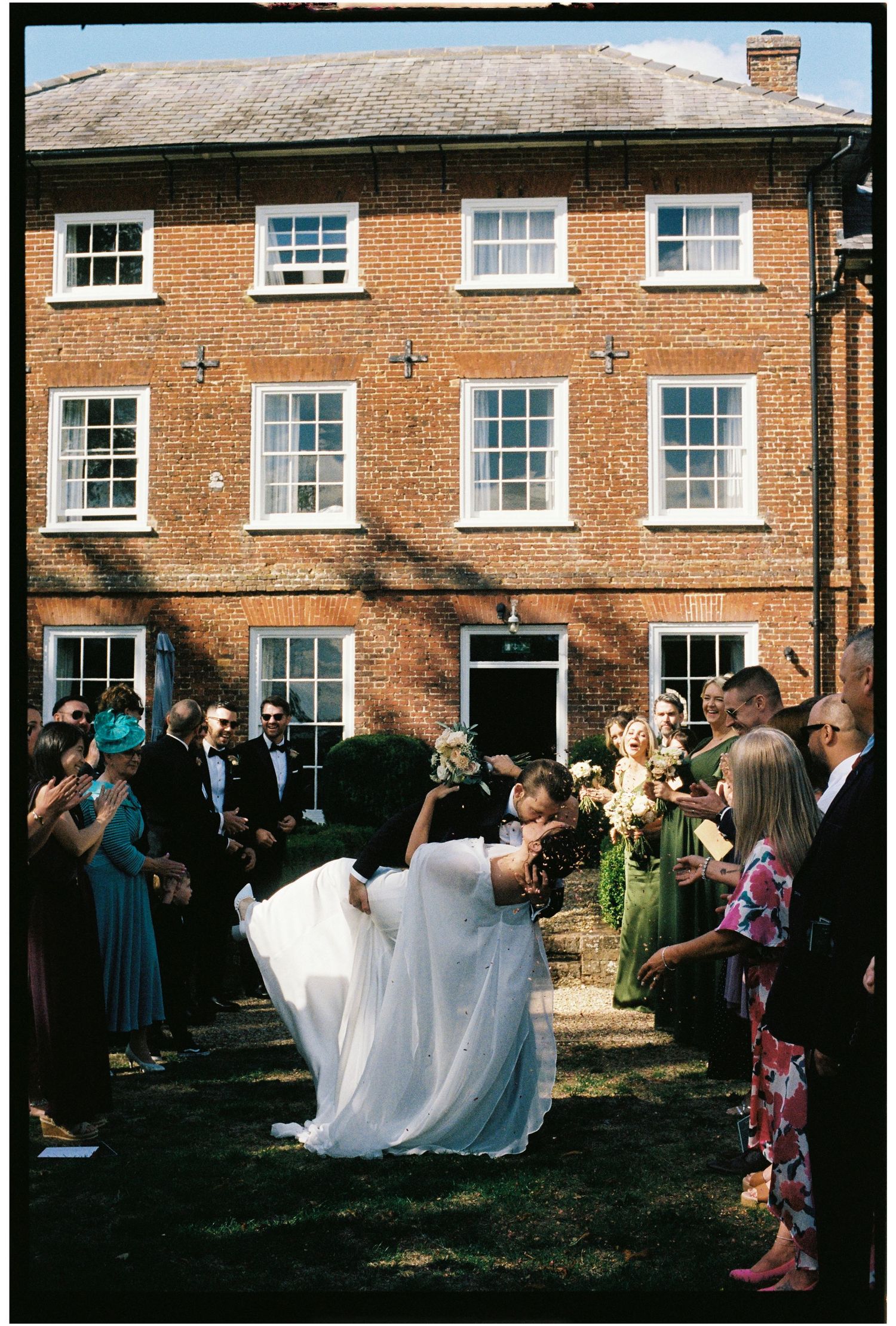 Groom dips bride outside elegant red brick wedding venue while guests cheer and applaud on a sunny day.