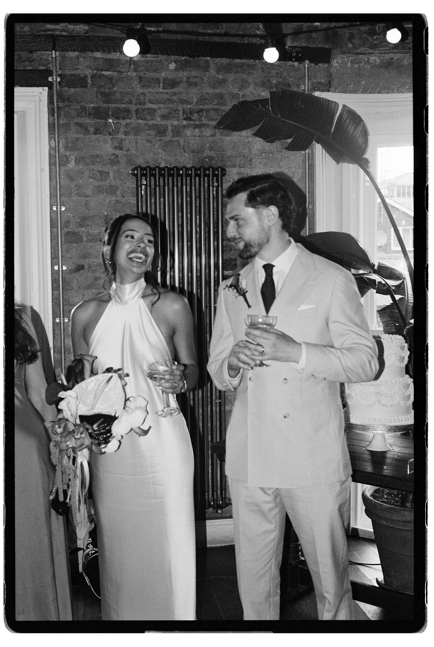 Black and white photo of laughing bride in halter gown and groom in suit holding drinks at reception.
