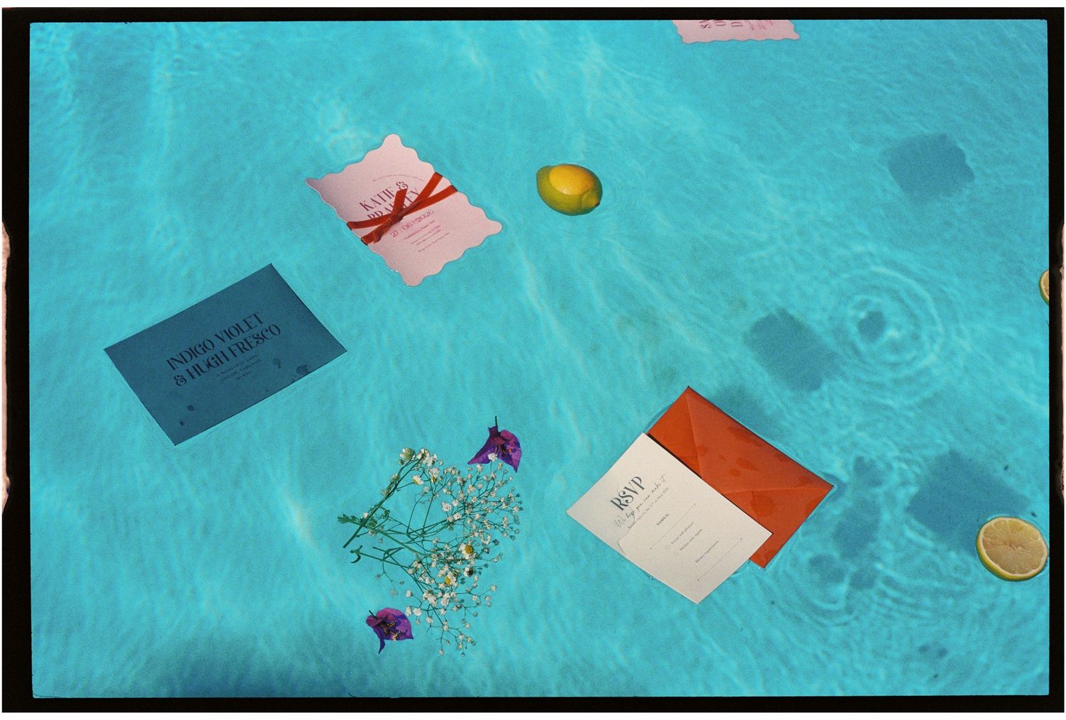 Colorful wedding invitations and citrus fruits floating in a bright turquoise swimming pool.