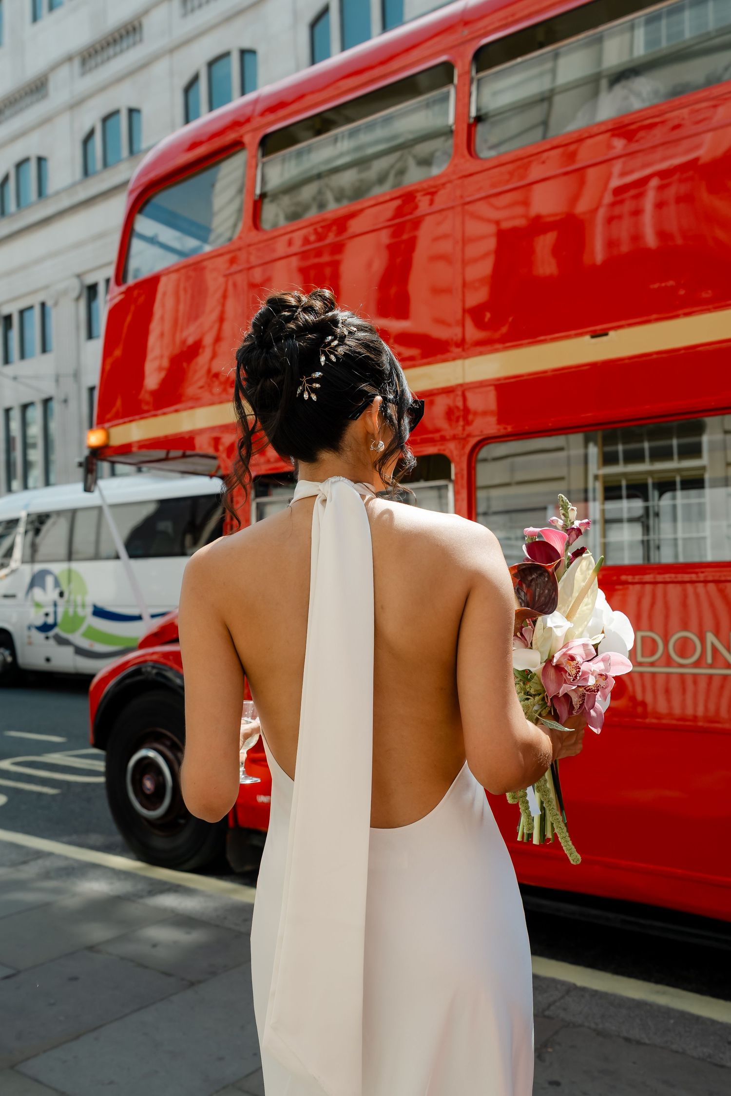 Bride in backless halter wedding dress holding bouquet standing before iconic red London double decker bus.