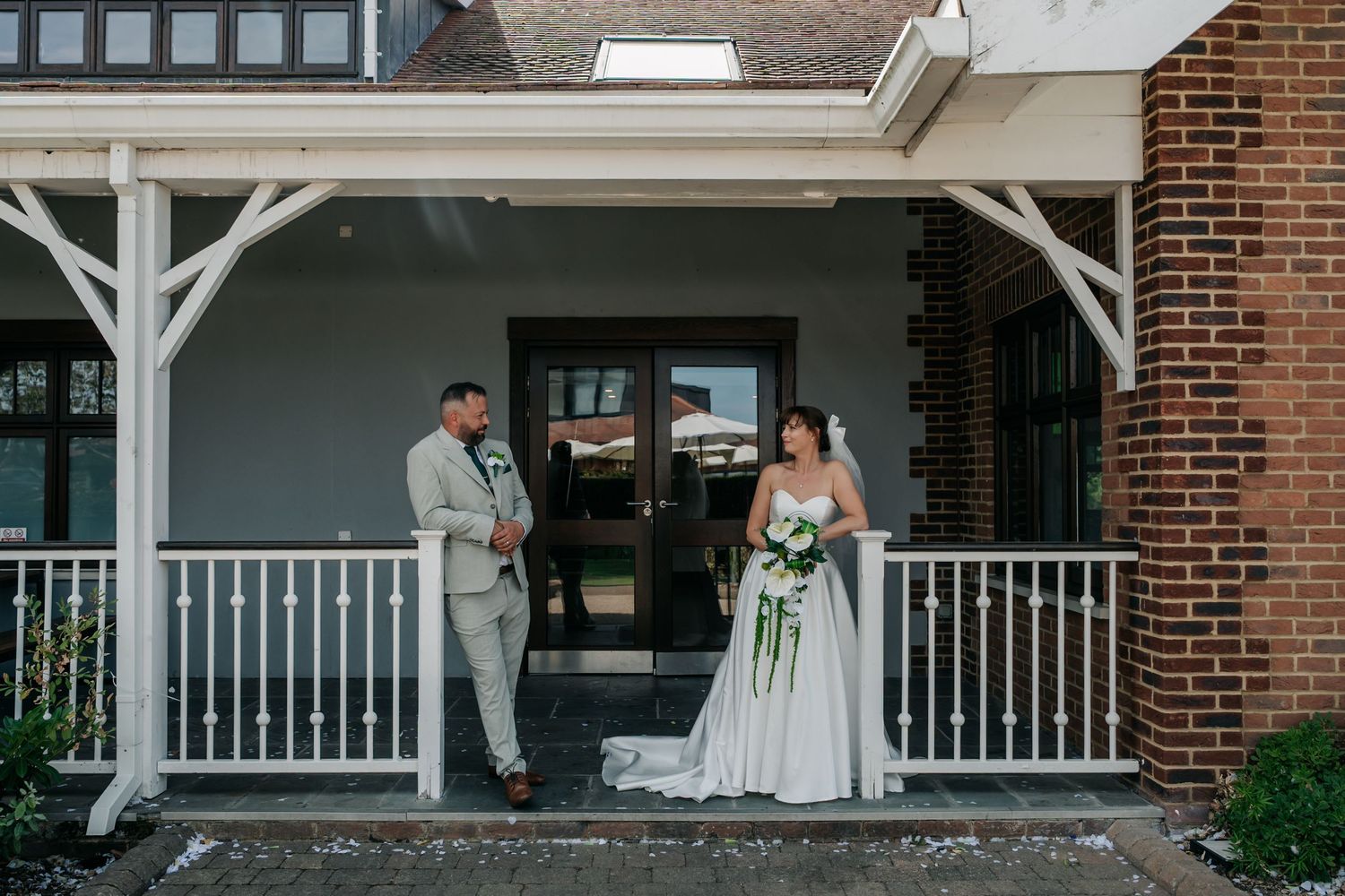 Bride and groom pose on a white porch of a brick building on their wedding day.