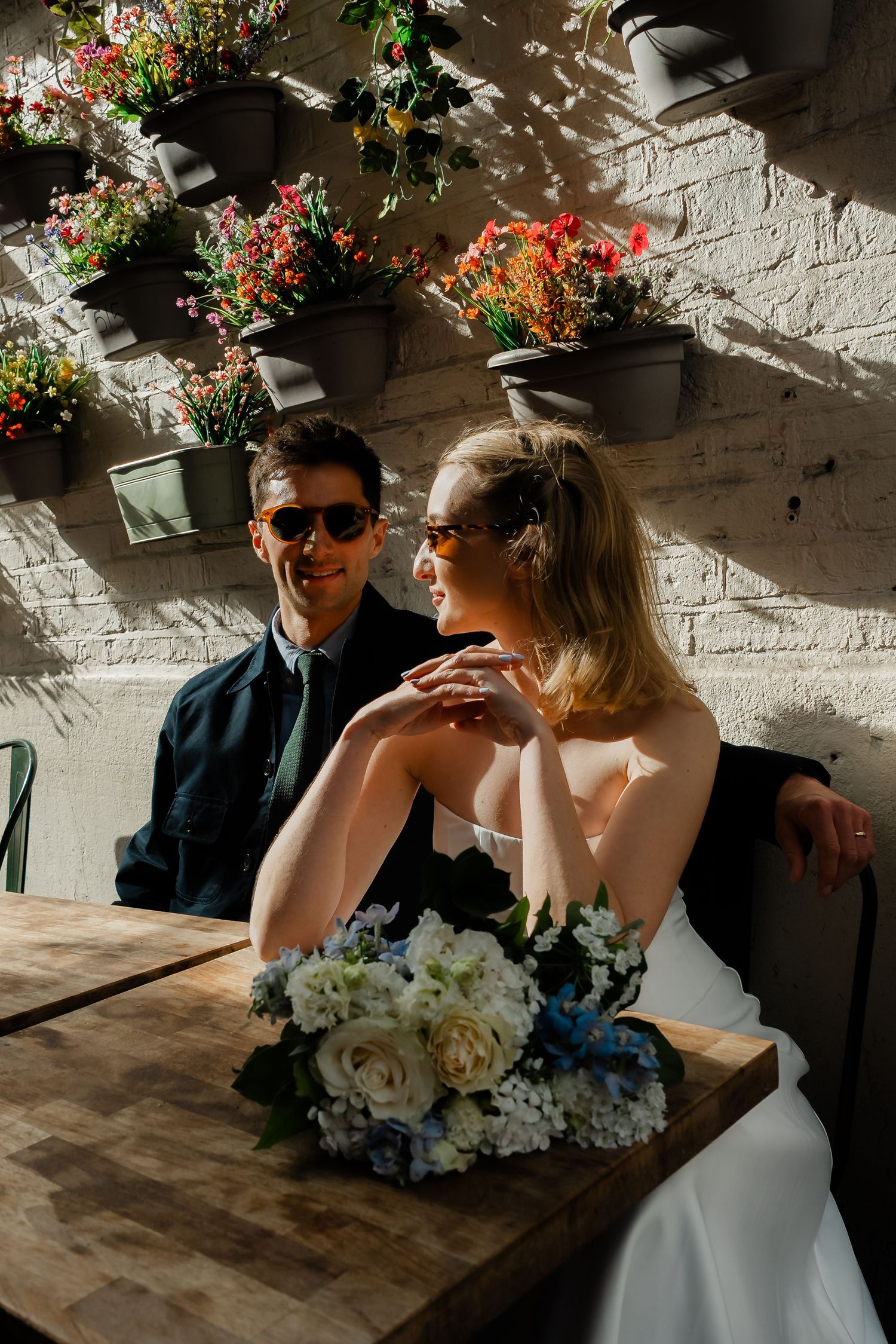 Bride and groom wearing sunglasses smiling at outdoor table with floral bouquet in sunlight.
