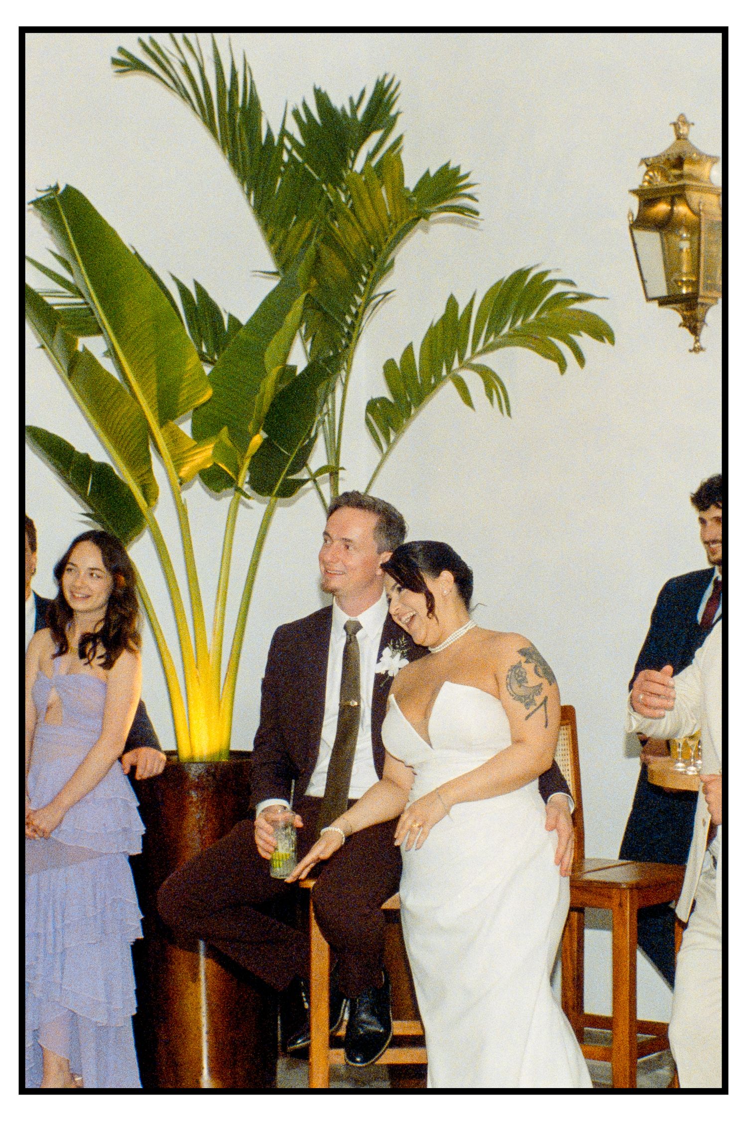 Bride and groom relax together at wedding reception surrounded by guests and tropical palm decor.