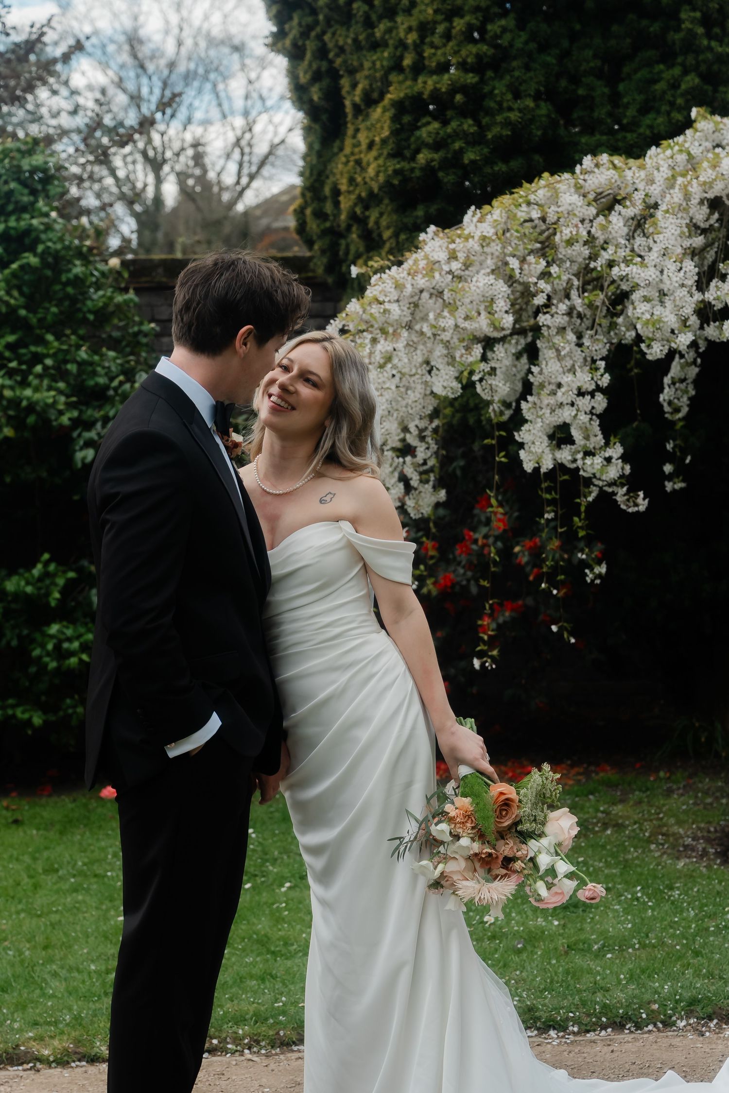 Bride in off-shoulder white gown and groom in black tuxedo sharing a romantic moment in garden.