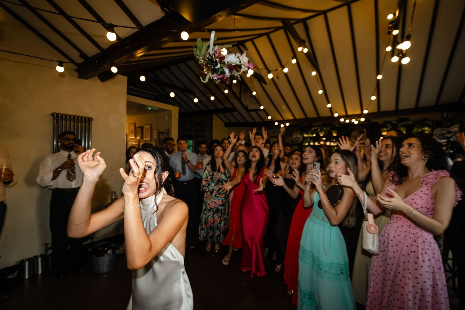 Bride tossing wedding bouquet to excited guests at indoor reception with string lights.