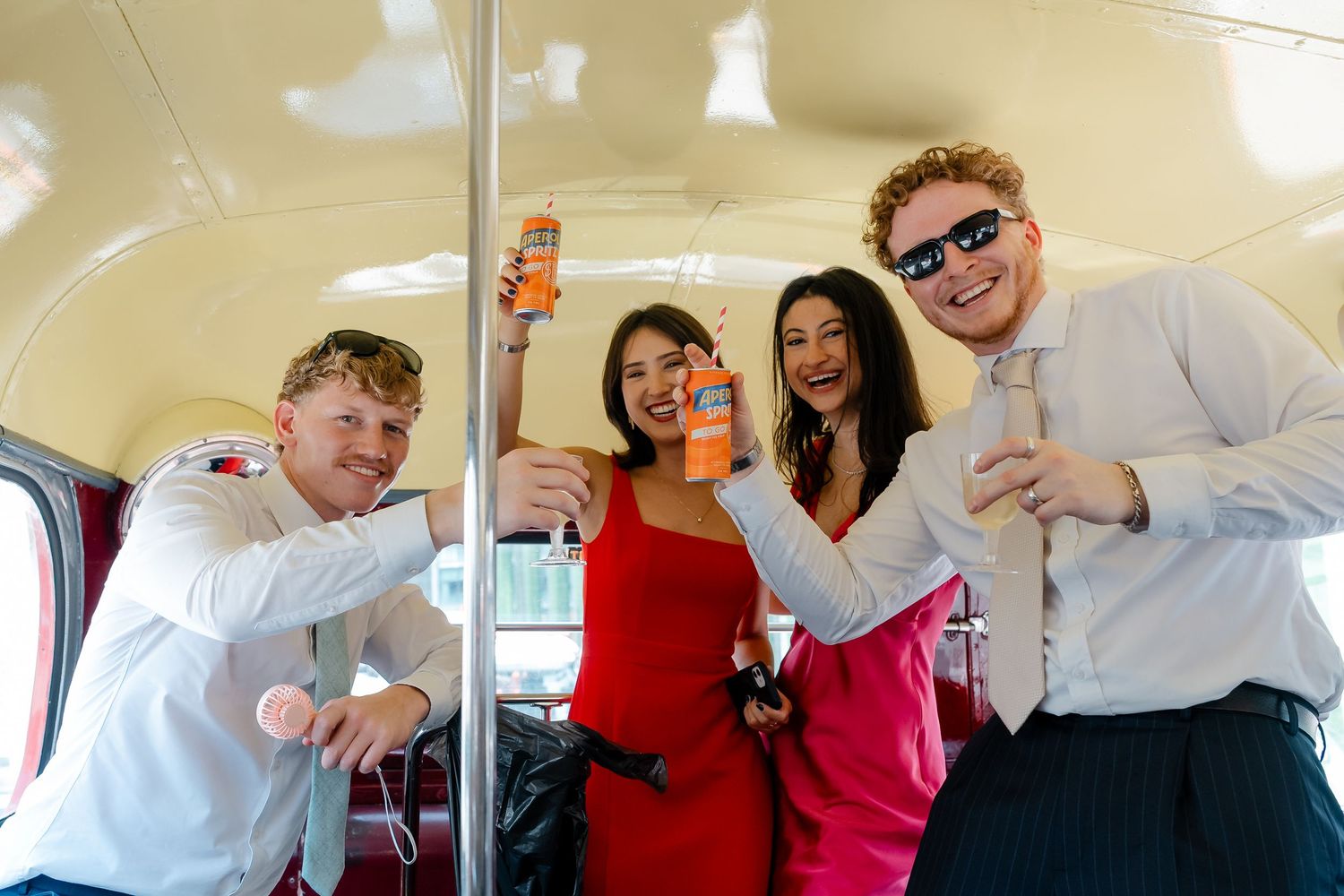 Four joyful wedding guests celebrating on a vintage bus, holding drinks and posing with confetti in festive attire.
