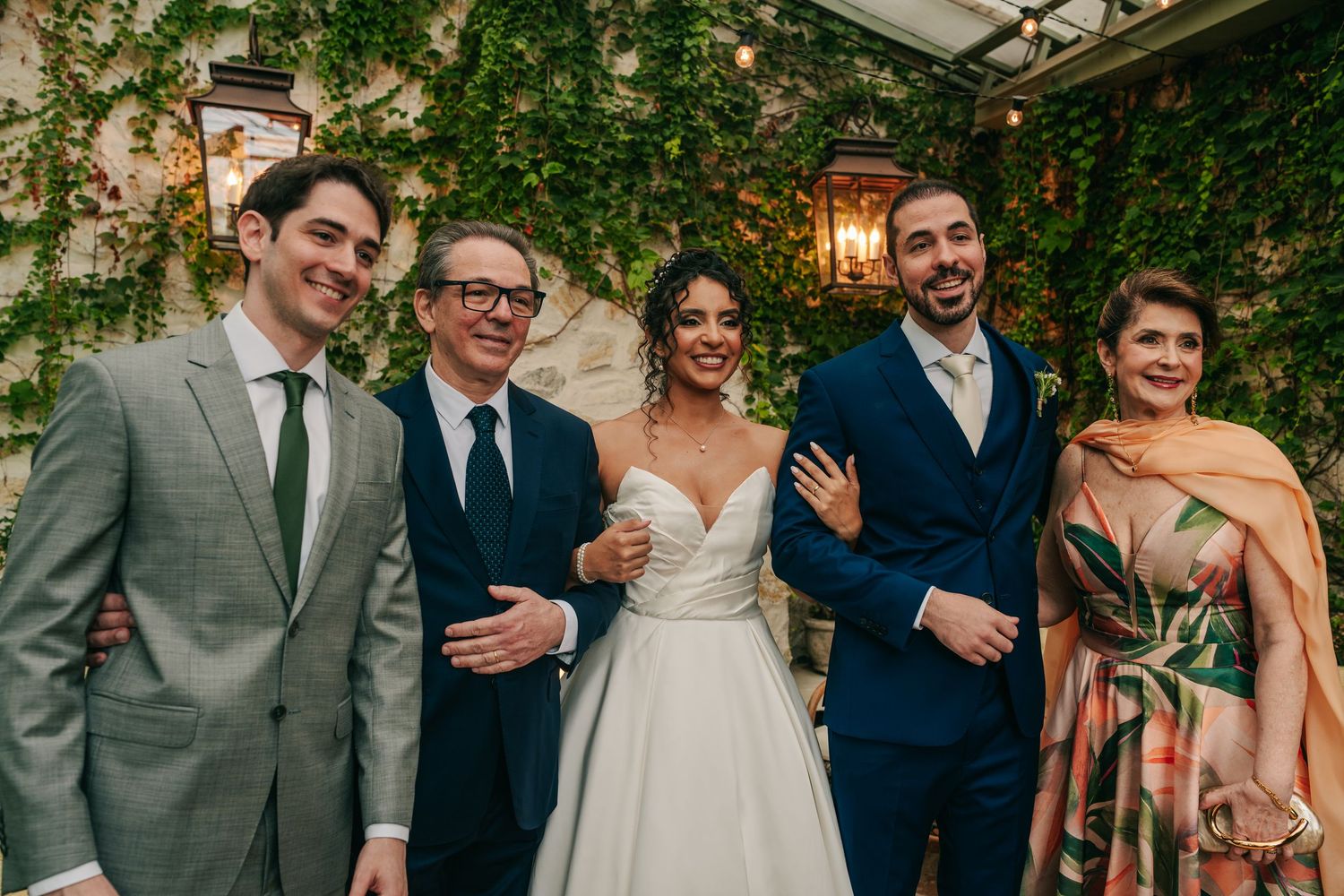 Newlywed couple smiles with family members in formal attire at elegant garden wedding venue.