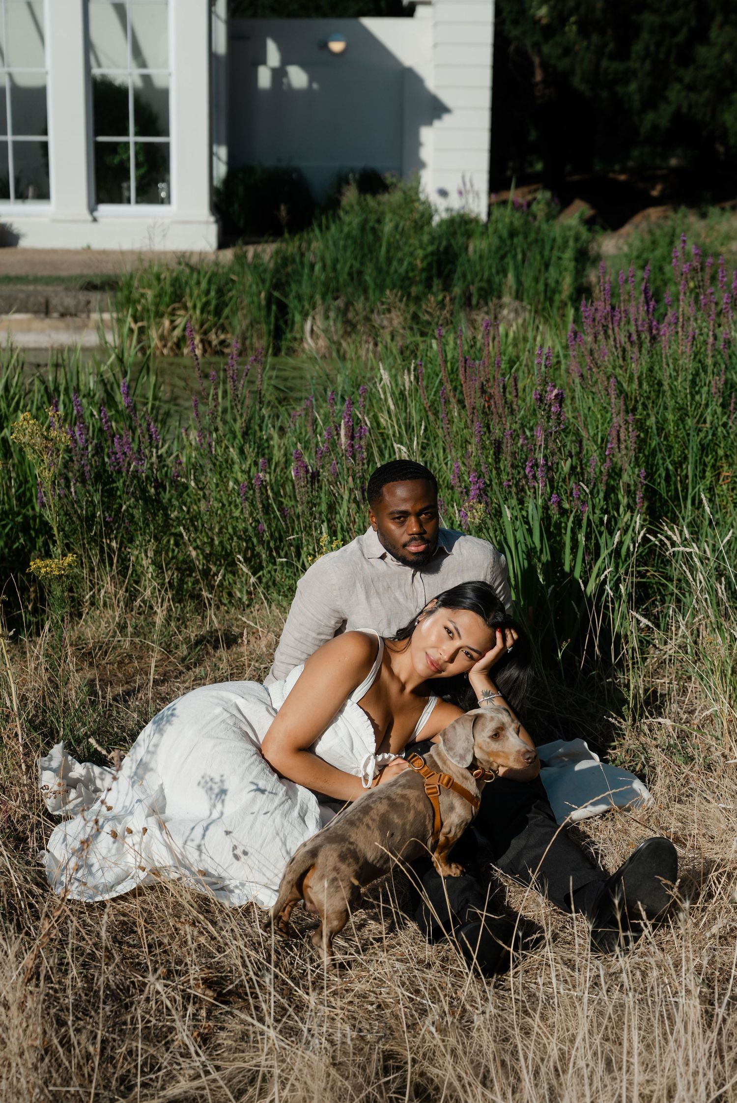 Couple relaxing in tall grass with a dog near a white building surrounded by lavender flowers.