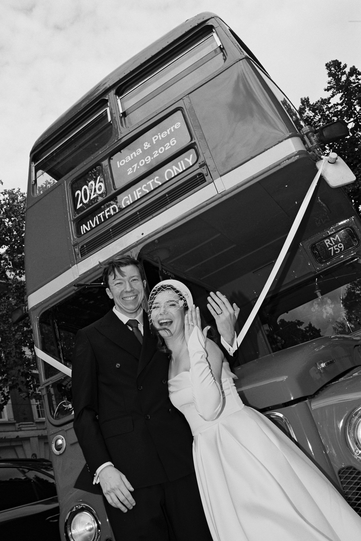 Black and white photo of a bride and groom laughing joyfully in front of a vintage double-decker wedding bus.