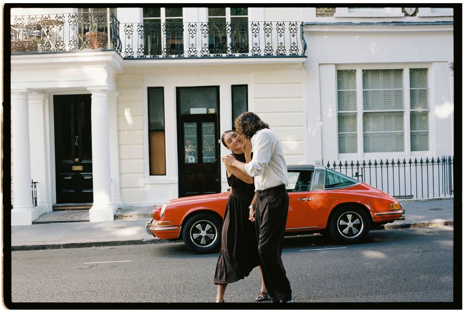 Couple embracing and kissing on a street in front of a classic orange Porsche 911 and white European townhouse.