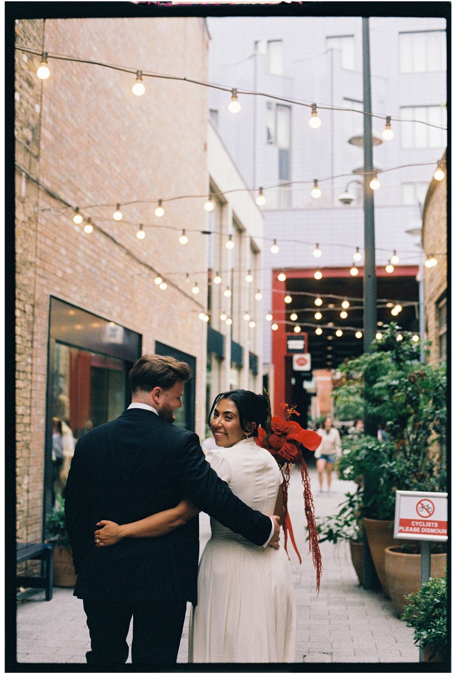 Bride and groom embrace in a charming brick alley adorned with string lights, bride holding a red bouquet.
