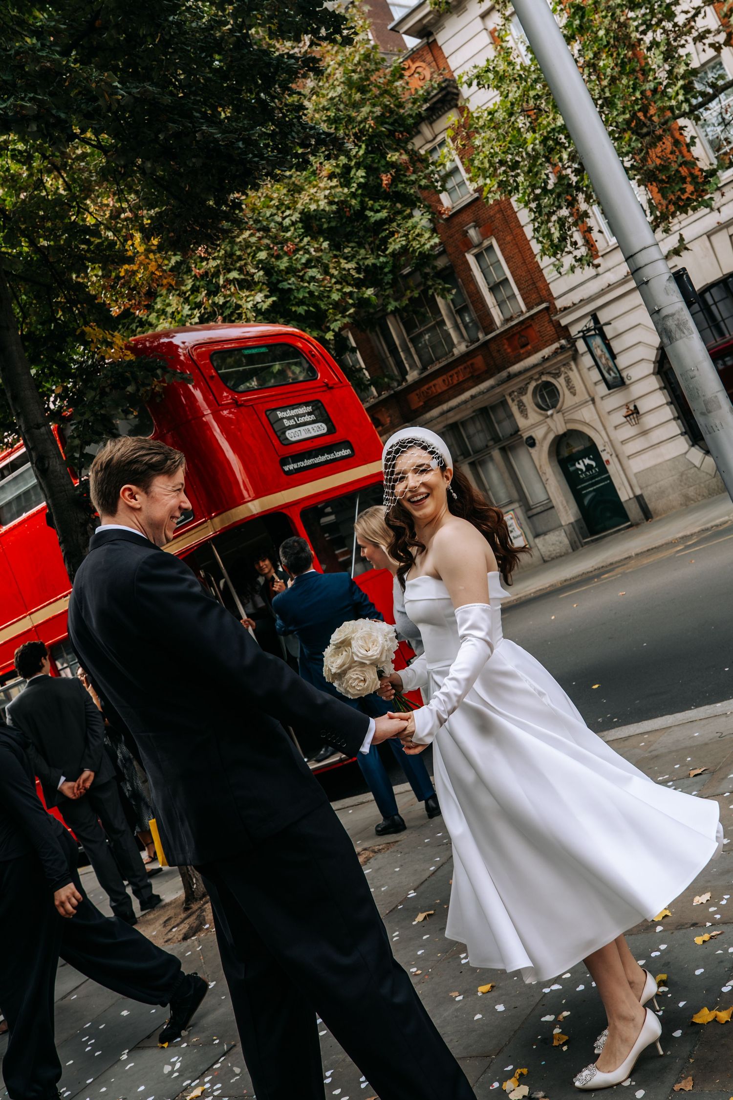 A bride and groom hold hands on a London street, with a iconic red double-decker bus behind them.