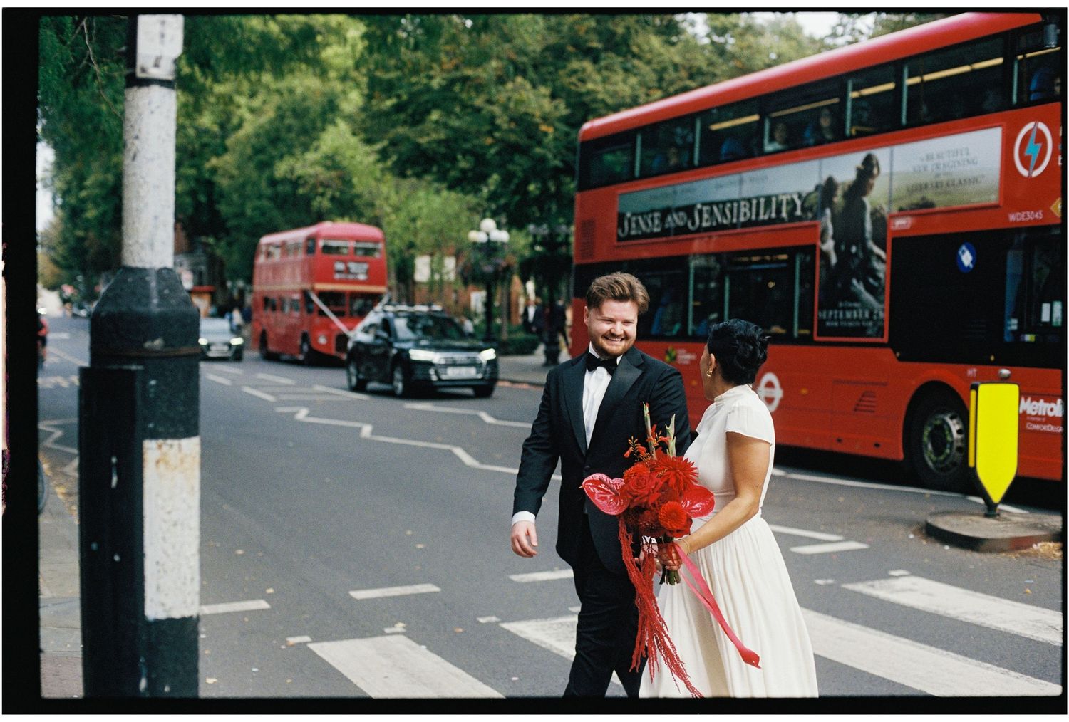 Newlywed couple crossing street in London with red double-decker buses, groom in tuxedo, bride holding red bouquet.