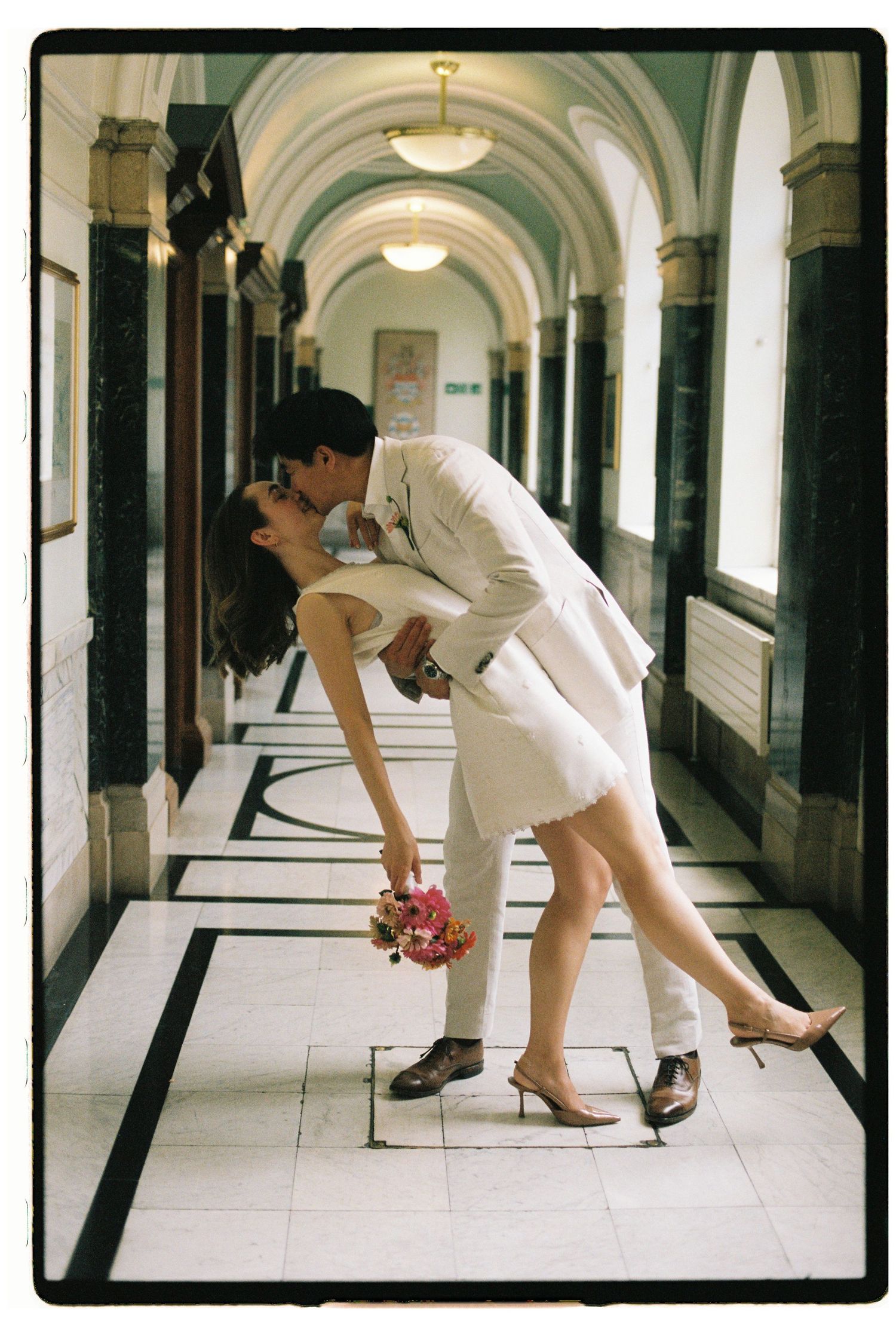 Couple sharing a romantic dip kiss in an elegant hallway, bride in white mini dress holding pink bouquet.