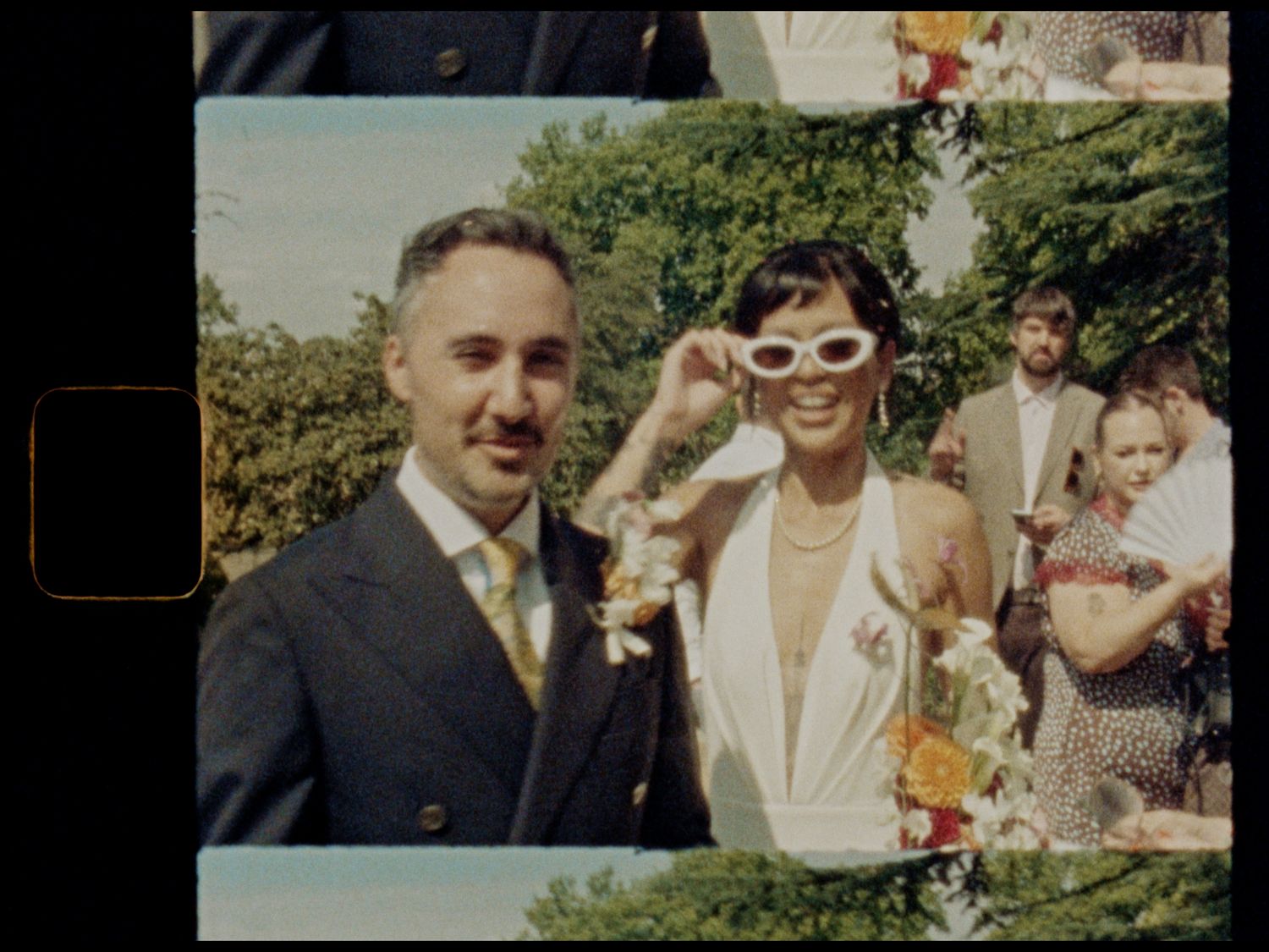 A couple poses together at an outdoor wedding, the bride wearing white sunglasses and gown, groom in navy suit.