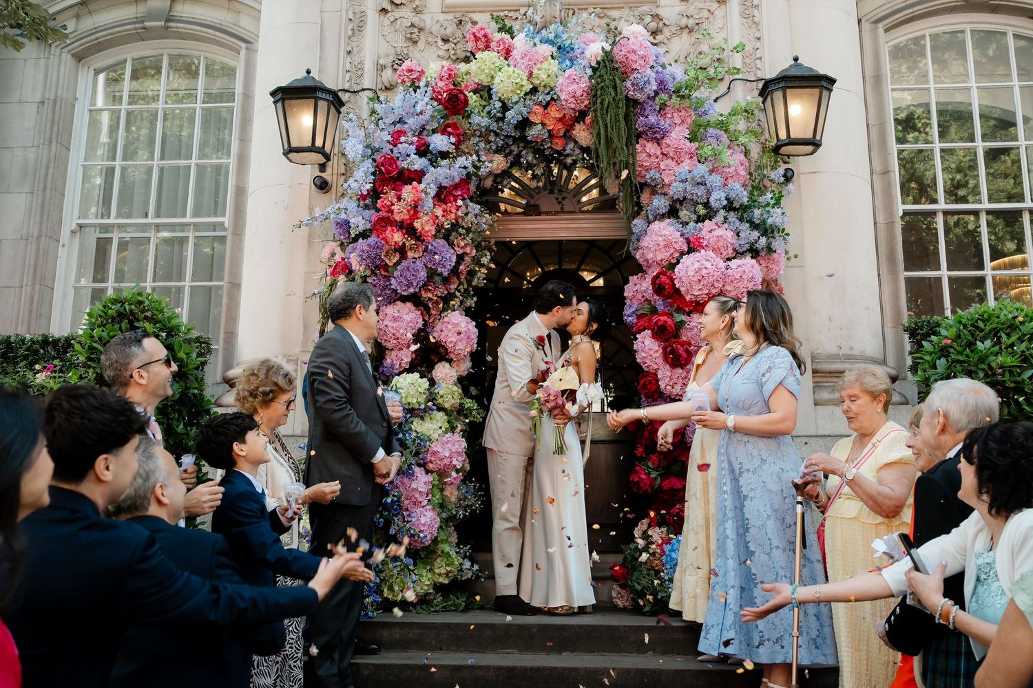 Couple kissing under colorful floral arch at wedding venue entrance.