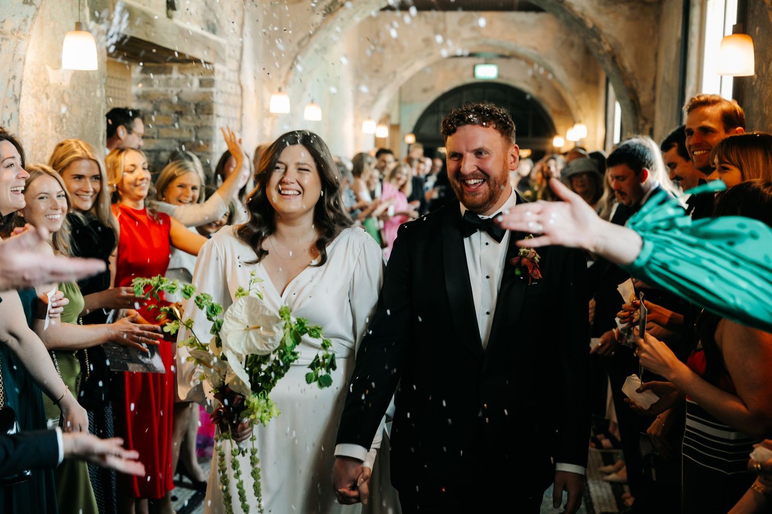 Bride and groom walk through confetti celebration in a stone archway venue.