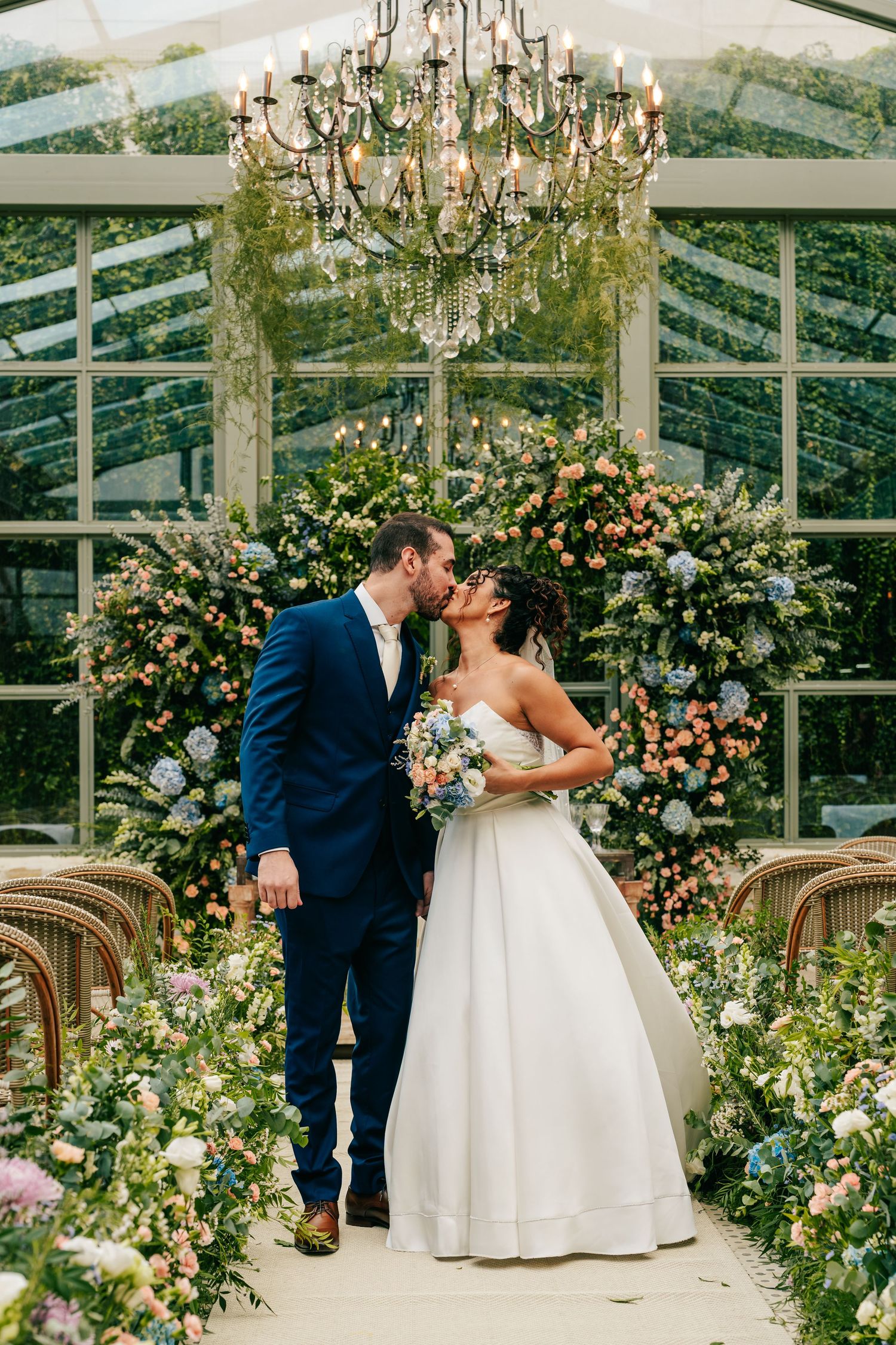 Newlyweds kiss at floral altar in glass greenhouse chapel with chandelier overhead.