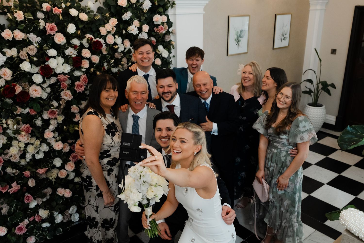 Bride in white dress takes selfie with wedding guests before elegant floral wall arrangement.