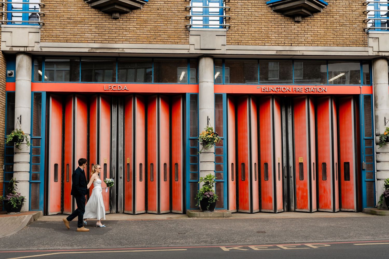 Bride and groom walking past vibrant red fire station doors on their wedding day.