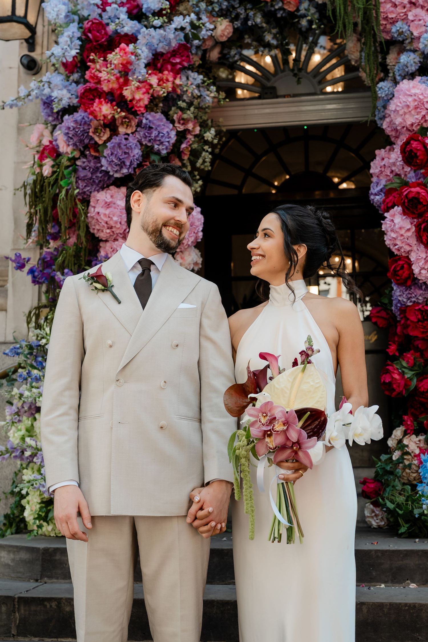 Smiling newlyweds holding hands before vibrant colorful floral arch, bride in halter dress with orchid bouquet.