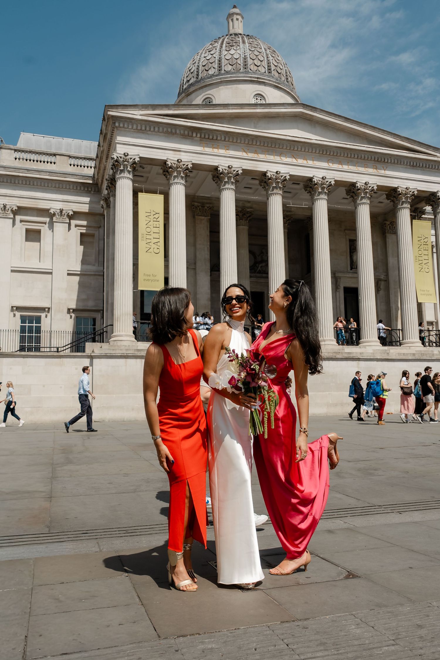 Bride in white halter gown with two bridesmaids in red and pink dresses outside the National Gallery London.