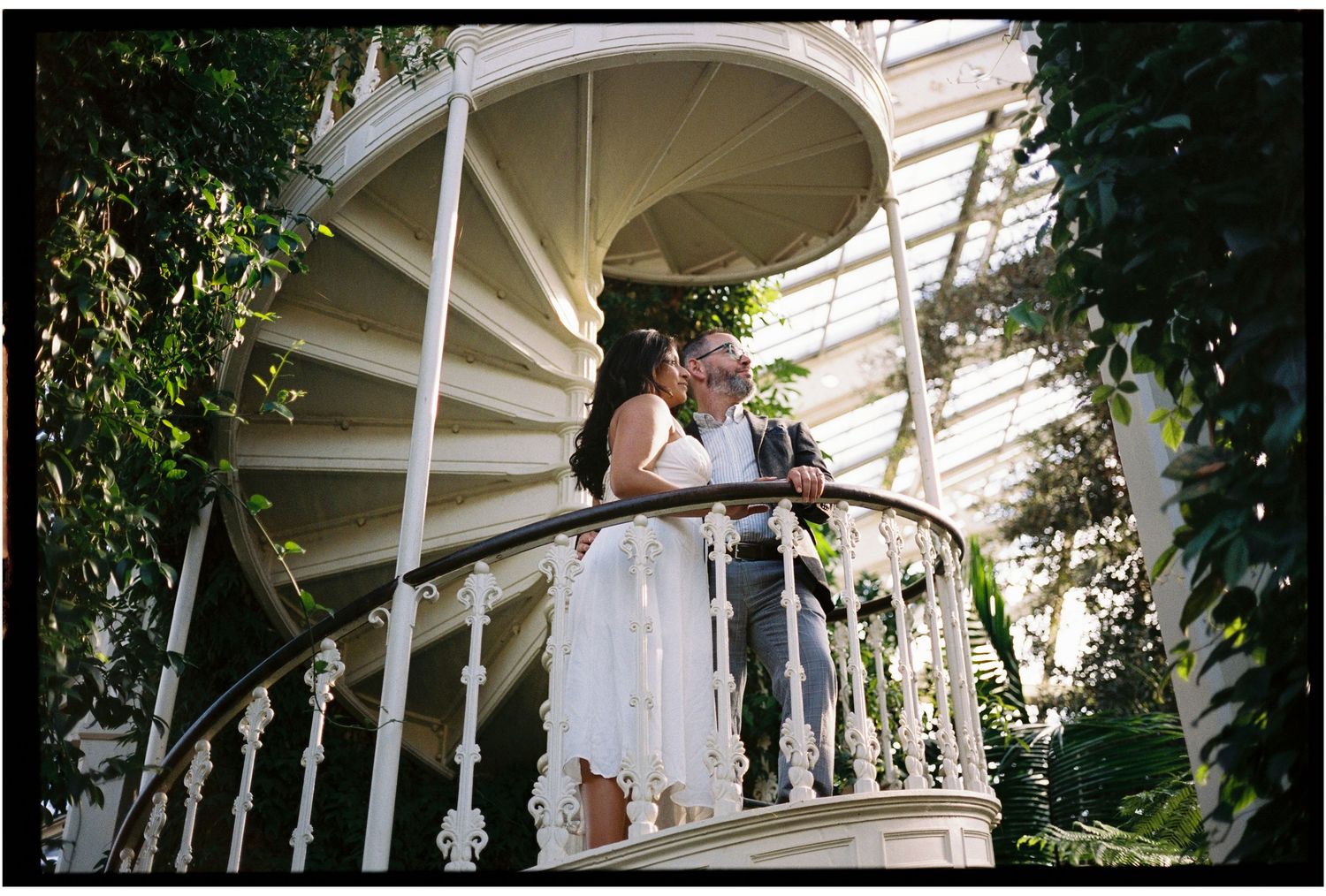 Couple in wedding attire embracing on ornate white spiral staircase balcony surrounded by lush green foliage.