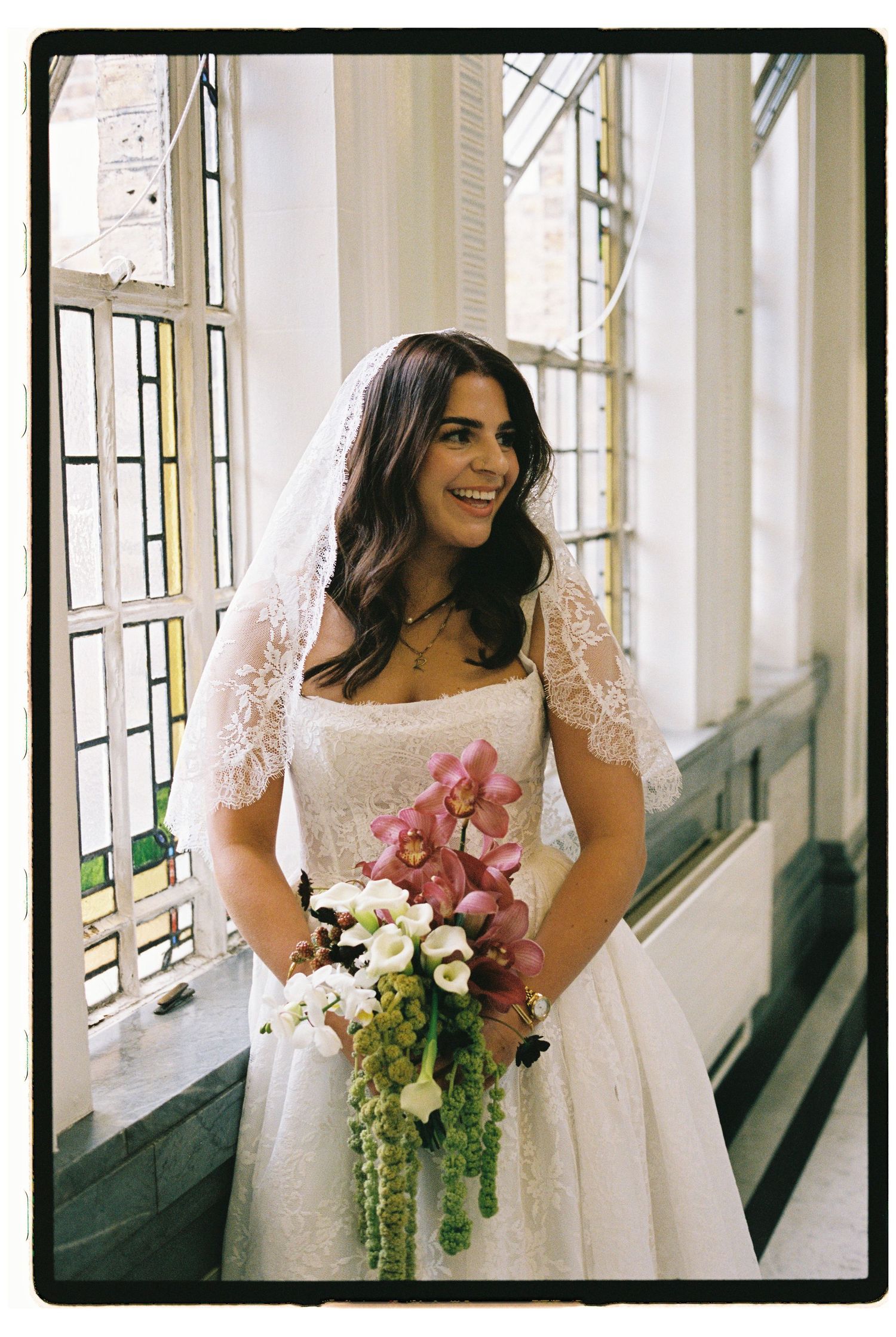 Smiling bride in lace wedding dress and veil holds pink orchid bouquet near stained glass windows.
