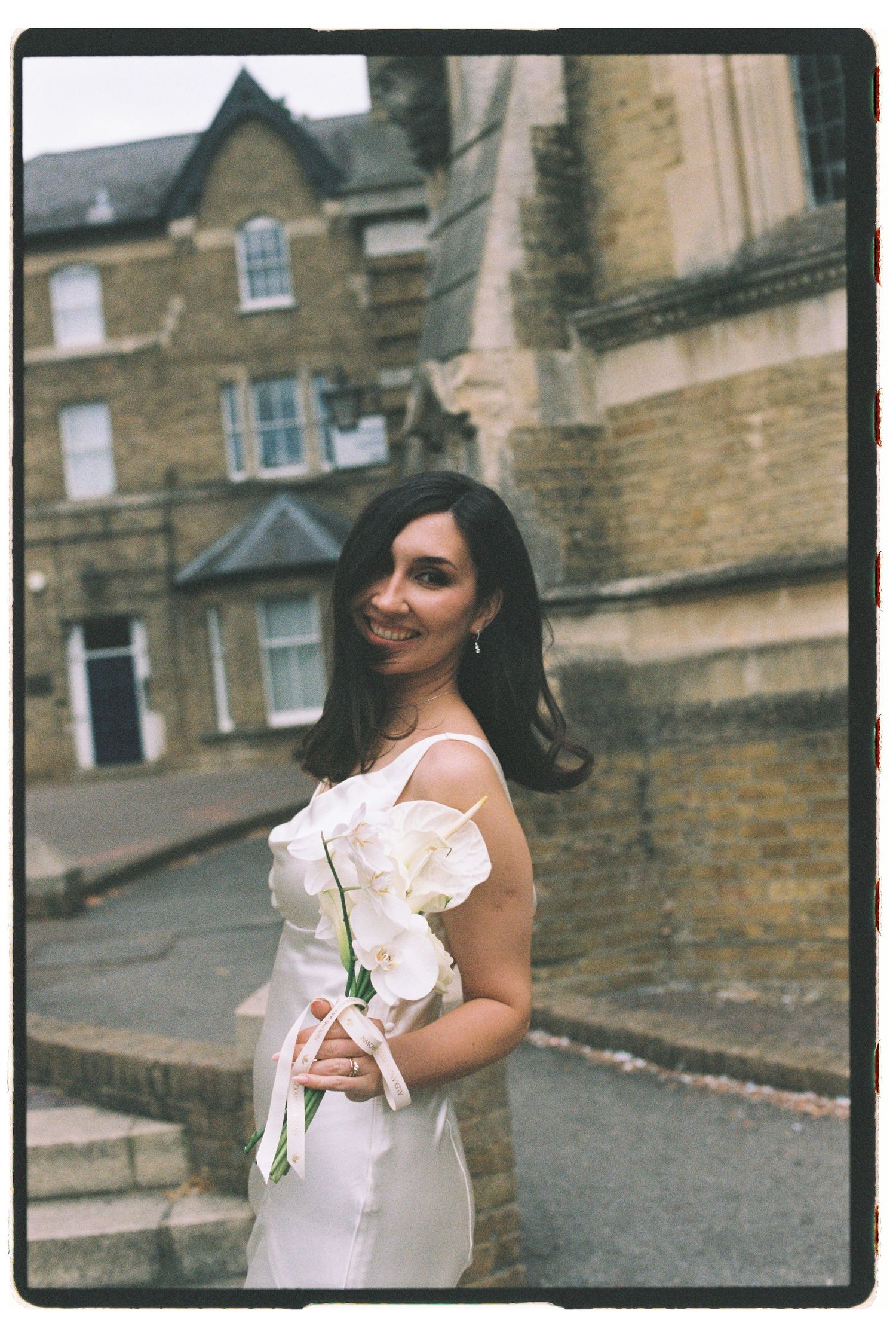 Smiling bride in white dress holding white orchids, posing on a cobblestone street with historic stone buildings.