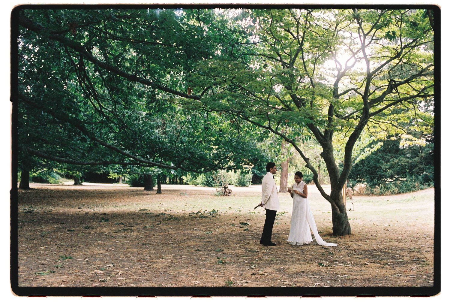 A bride and groom hold hands under lush green trees in a scenic outdoor park wedding photo.