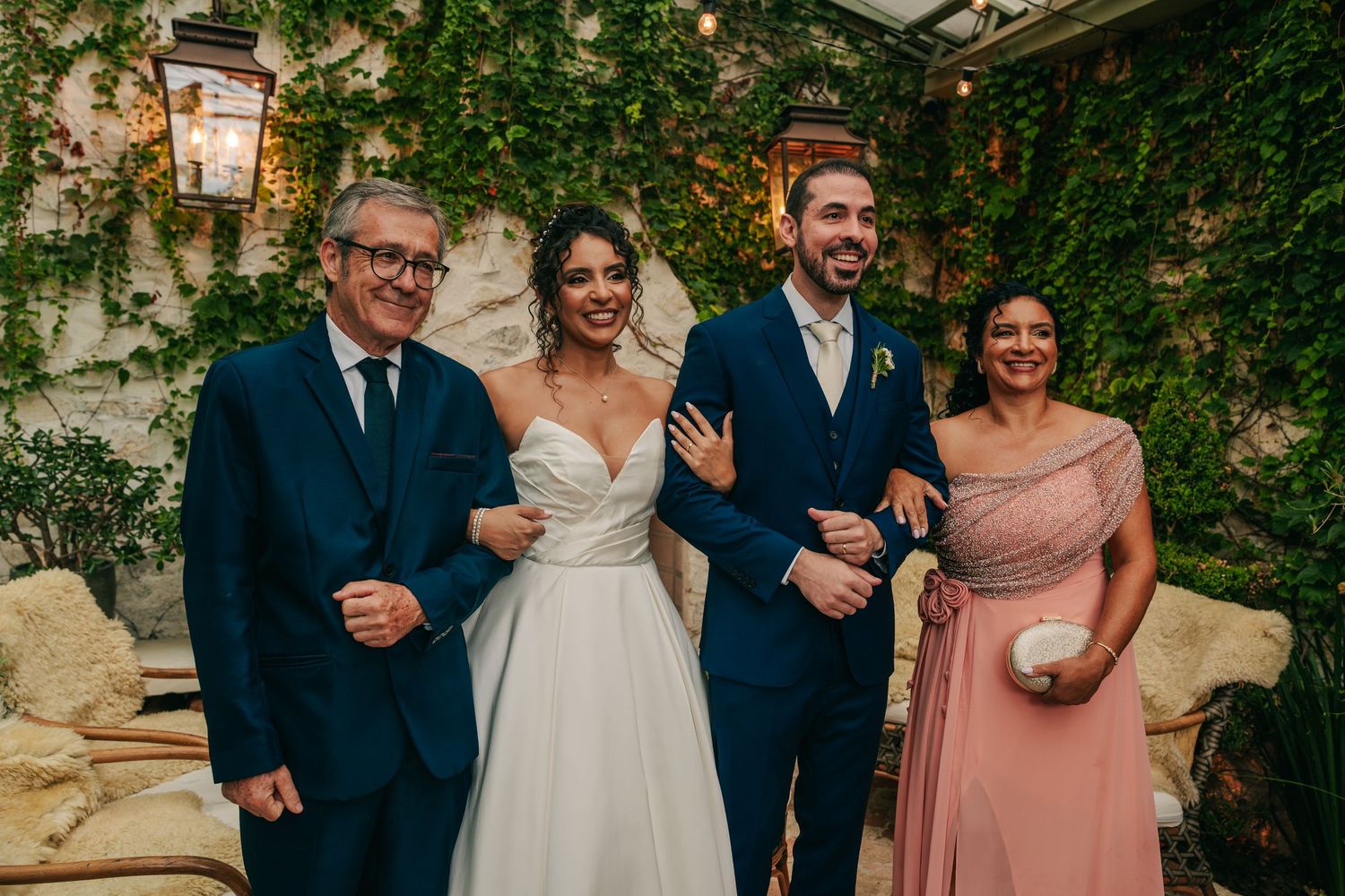 Bride and groom pose with parents in formal attire against lush ivy wall at wedding.