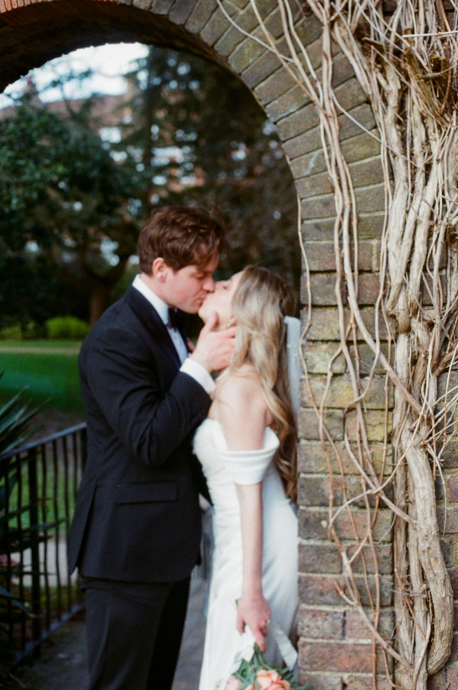 Bride and groom kissing beneath rustic brick archway covered in climbing vines at elegant wedding venue.