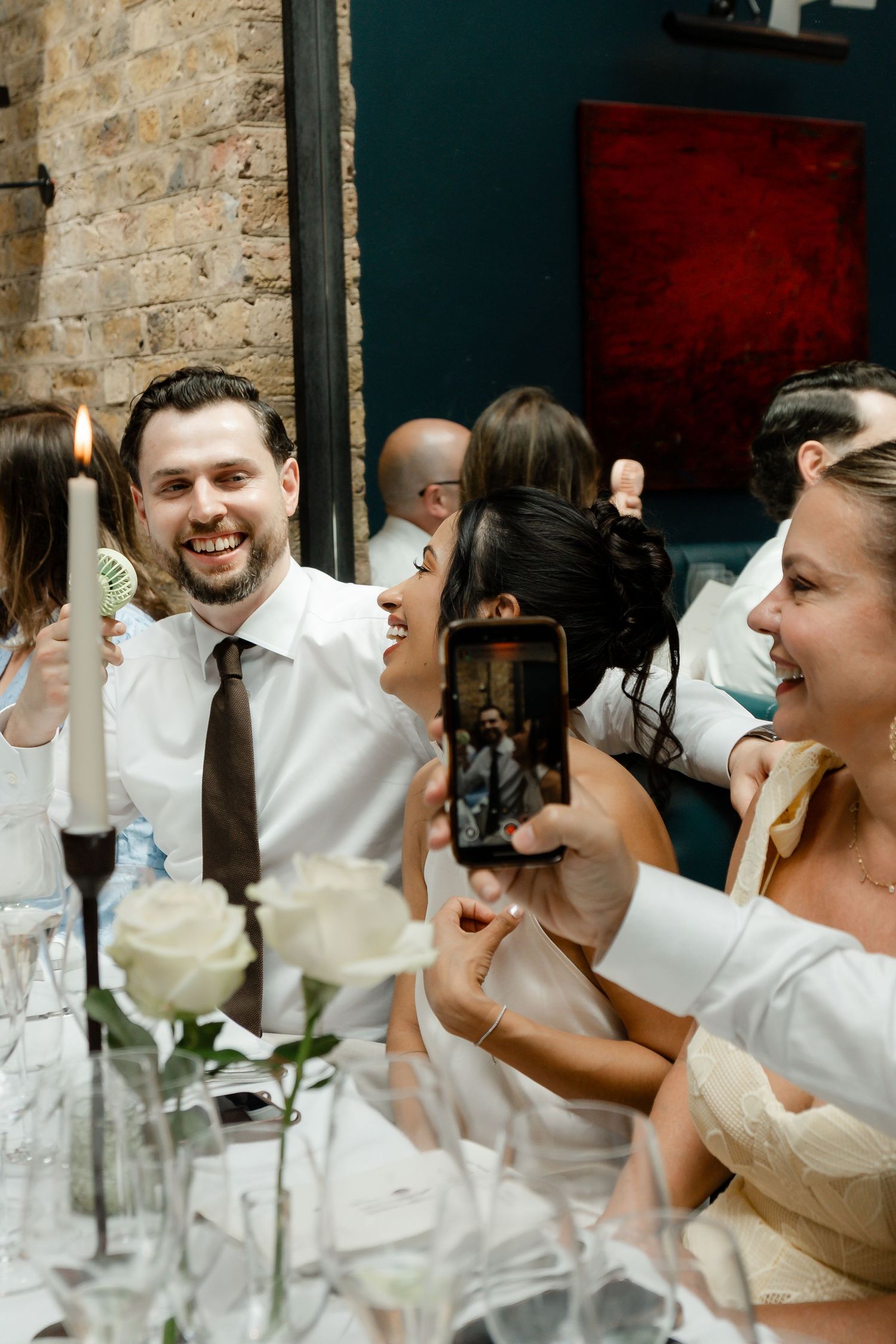 Groom holding a microphone and candle while laughing with guests during a lively wedding reception.