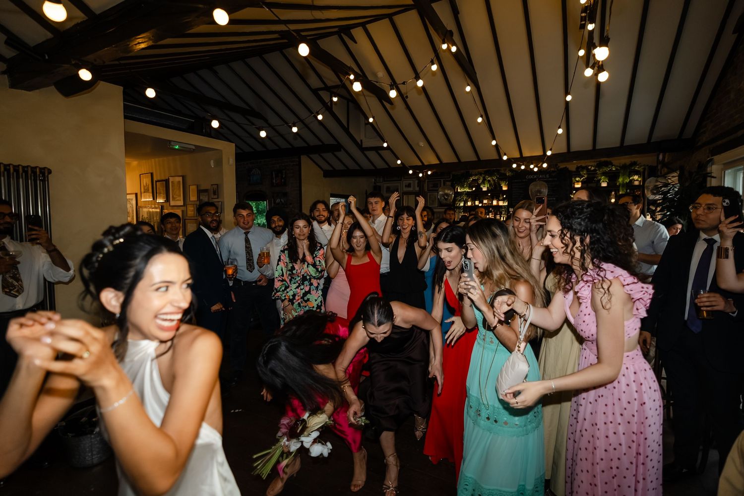 Wedding guests eagerly reaching for the bouquet toss at a lively indoor reception celebration.
