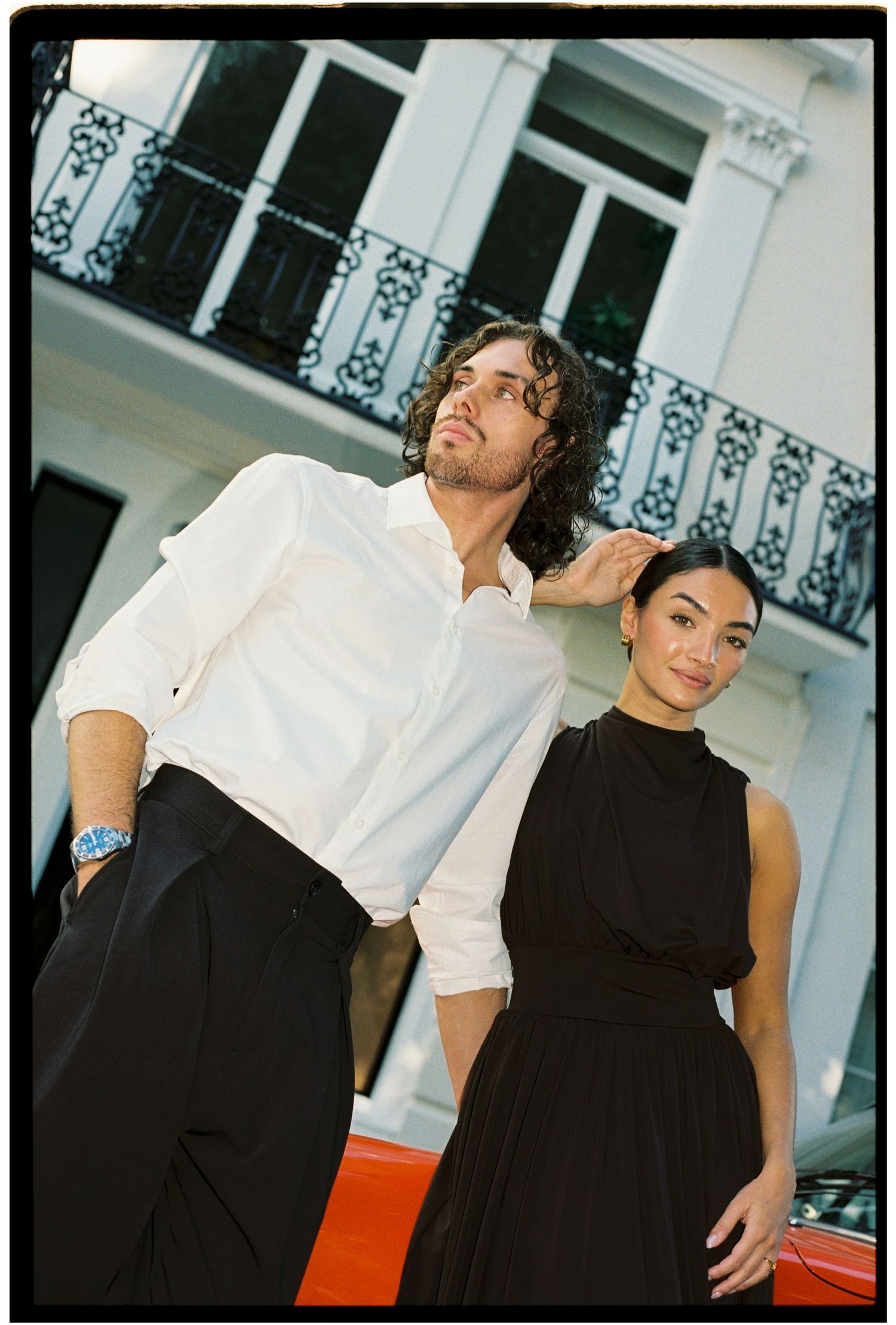 A man in white shirt and woman in black dress pose elegantly in front of a white building with balcony.