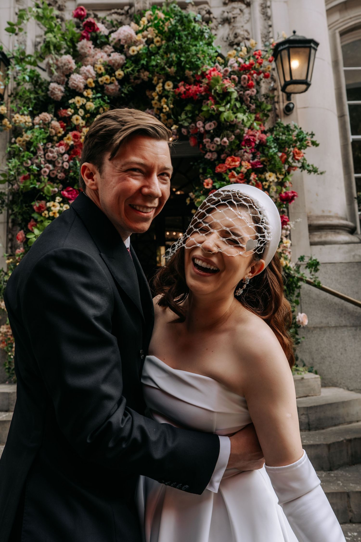 Joyful bride in white strapless gown and birdcage veil embraced by groom in black suit outside floral venue.