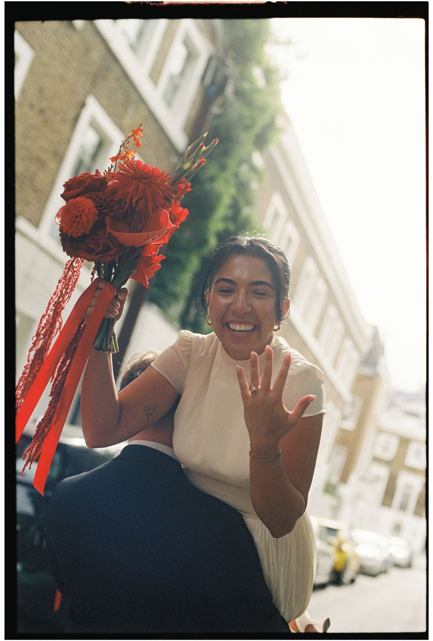 Joyful bride shows off wedding ring while holding a vibrant red floral bouquet on a sunny street.